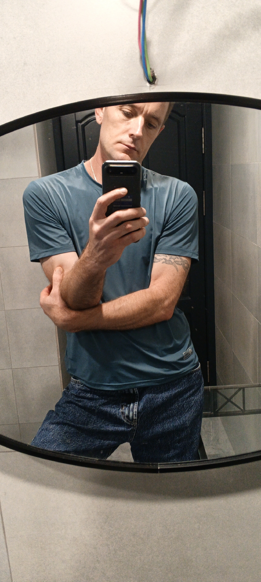 A man in a teal t-shirt and blue jeans is taking a mirror selfie in a bathroom. He is holding a black smartphone in his right hand, with his left arm crossed over his chest. The mirror is oval-shaped with a black frame and is positioned on a grey tiled wall. In the background, a dark doorway and some colorful electrical wires are visible. The lighting appears to be artificial, creating shadows on the wall. The man's expression is neutral as he looks at his reflection. The location is not specified in the image, and therefore the city of Tashkent, Uzbekistan, cannot be determined.