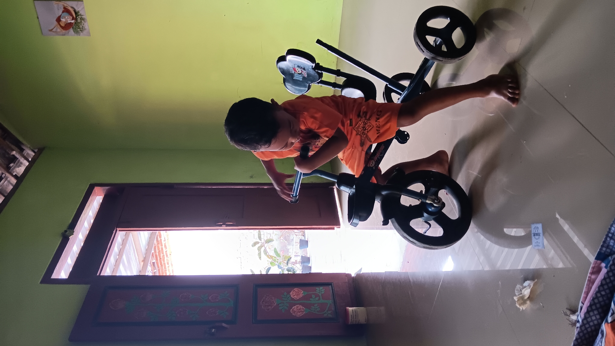 A young boy in an orange t-shirt and shorts sits on a black tricycle indoors. He is facing left and leaning forward, holding the handlebars with both hands. The tricycle has a front basket and two rear wheels. The boy's feet are bare. The scene is set in a room with lime green walls. To the left, a dark red door is open, revealing an outdoor area with plants in pots and a corrugated roof. Sunlight streams through the doorway, casting a shadow from the tricycle and the boy onto the shiny tiled floor. A small piece of crumpled white paper and a small white rectangular object are on the floor in front of the tricycle.