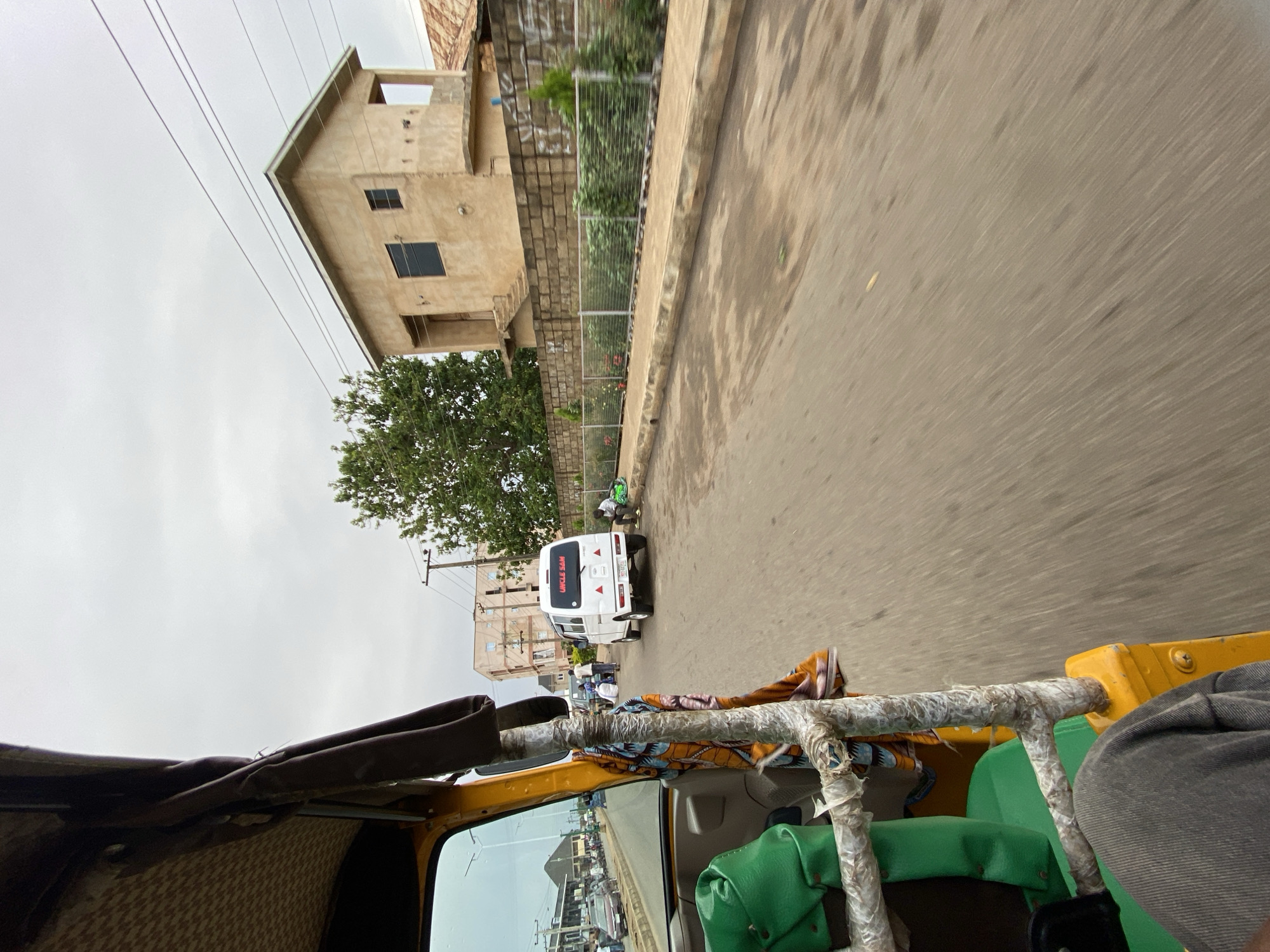 The image shows a view from inside a yellow vehicle, likely an auto rickshaw, moving along a paved road in Bukuru, Nigeria. The road appears wet, indicating recent rain. A white van with the name "Uncle Sam" is ahead in the same direction. A tan, two-story building is on the left side of the road, and a tree and a partially visible fence run along the roadside. Overhead power lines are visible against a grey sky. There are a few individuals standing on the side of the road to the left, near the white van, and one more on the right side behind the van. The interior of the auto rickshaw is visible, including a green seat, metal frame, and brown roof. A person's arm and shoulder are partially visible on the right.