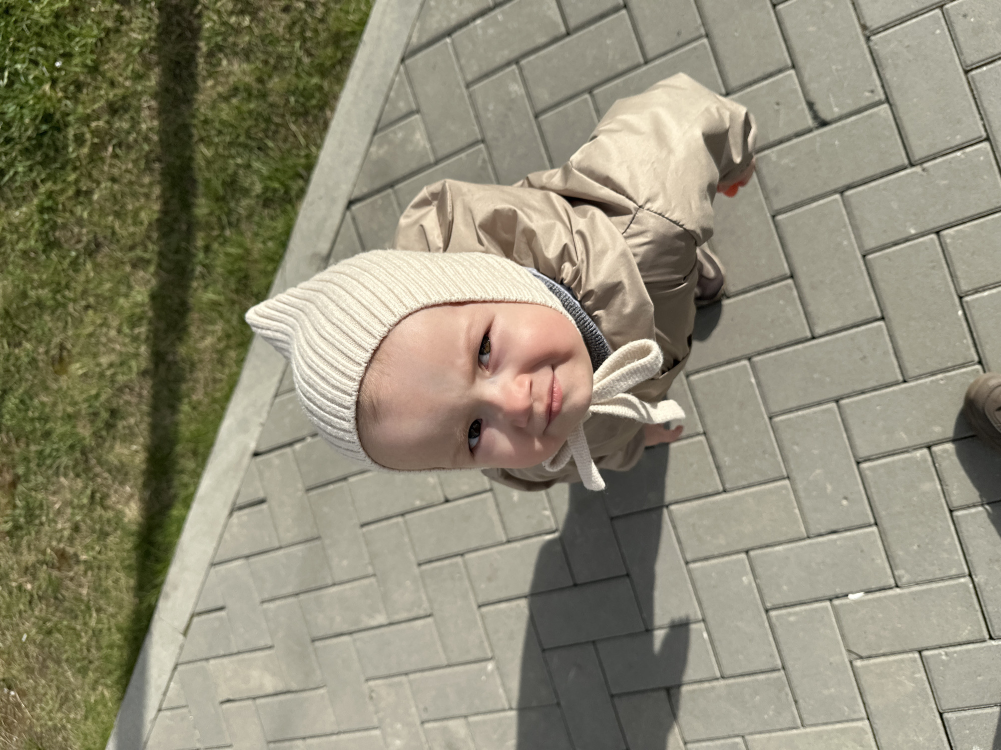 A young child, appearing to be a baby or toddler, is the central subject of the image, captured from a high angle looking down. The child wears a light cream-colored, ribbed bonnet with ties under the chin and a light tan or beige puffer-style jacket. They are looking directly up at the camera with a gentle, curious smile and wide, dark eyes.
The child is standing on a grey interlocking paver pathway, laid out in a herringbone pattern. To the left of the pathway, a strip of green grass is visible. Strong, defined shadows are cast across both the grass and the pavement, indicating bright sunlight and suggesting a clear, sunny day. A long, thin shadow, possibly from a pole or similar object, stretches across the grassy area. Another shadow, likely from the adult taking the picture, falls across the pavement near the child. On the far right edge of the image, part of an adult's dark brown shoe is visible.
The scene takes place outdoors, possibly in a park or pedestrian area, under bright daytime conditions. No visible text is present in the image. The location context for this scene is Yekaterinburg, Russia.