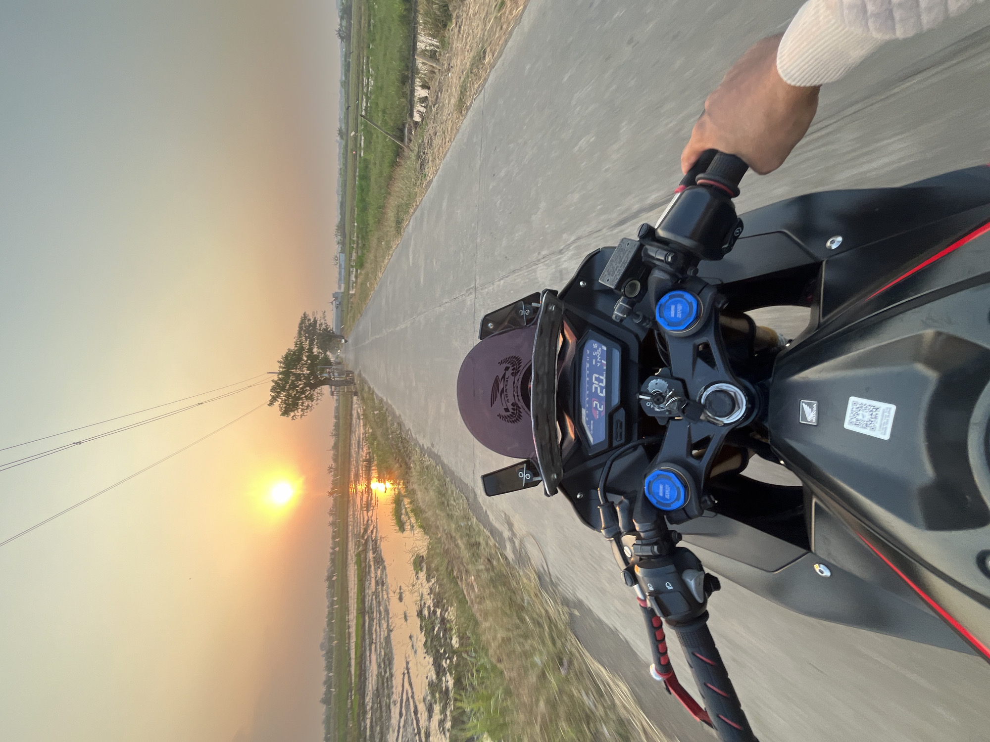 A first-person perspective shot captures a motorcycle rider cruising down a paved road during sunset in Pecangaan, Indonesia. The sun, a vibrant orange orb, hangs low in the sky, casting a warm glow over the surrounding rice paddies, which reflect the golden light. Power lines stretch across the horizon, disappearing towards distant trees. The motorcycle's handlebars, digital dashboard displaying "2 20.3", and part of the rider's arm are visible in the foreground. The landscape is characterized by flat terrain, with green fields and bodies of water suggesting agricultural land. The scene evokes a sense of peaceful travel through the Indonesian countryside at dusk.