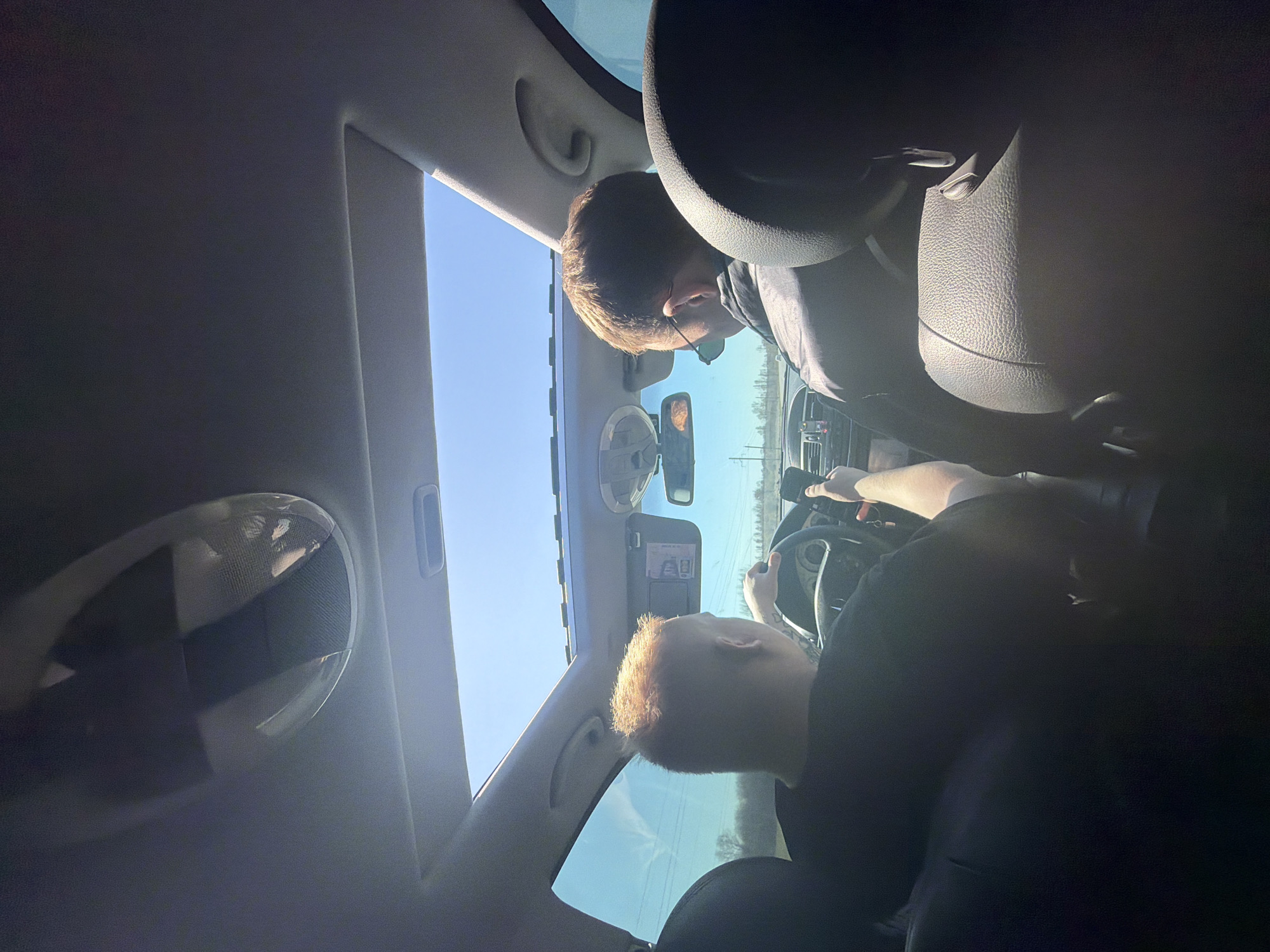 A low-angle photograph taken from within a car shows two individuals. One person is driving, their hands on the steering wheel. Another person is in the passenger seat. The car's sunroof is open, revealing a clear blue sky. A rearview mirror is visible. The location is inferred to be Гремяча, Russia based on user query.