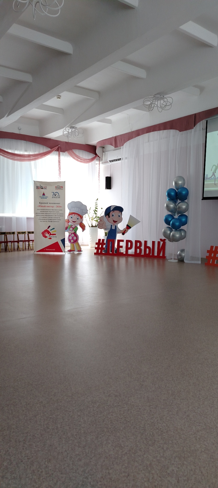 The image depicts an indoor setting likely in Chaikovsky, Russia, decorated for an event. A banner reads "Krasvoi championship "Young Master-2026". Two cartoon characters, one dressed as a chef and the other as a construction worker, stand next to the banner. Red lettering spelling "#ПЕРВЫЙ" is prominently displayed. A balloon arrangement of blue and silver balloons is positioned on the right side of the scene. The room has a light-colored floor and white ceiling with distinctive architectural details. Chairs are visible in the background, suggesting an event setup. White sheer curtains and a projector screen are also present.