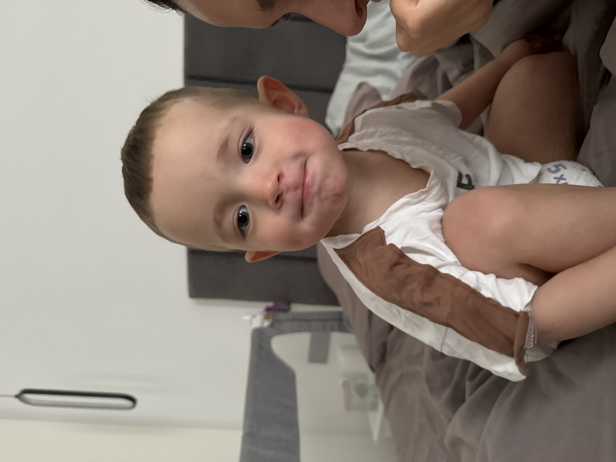 A young child with light brown hair and brown eyes is lying on a bed with gray bedding. The child is wearing a white shirt with brown trim. Part of a person's face is visible in the top of the frame, close to the child's face. The child's legs are visible, and they appear to be wearing a diaper. In the background, there is a bedside table with items on it, and a white wall. No landmarks indicating the city of Odintsovo, Russia are visible in the image.