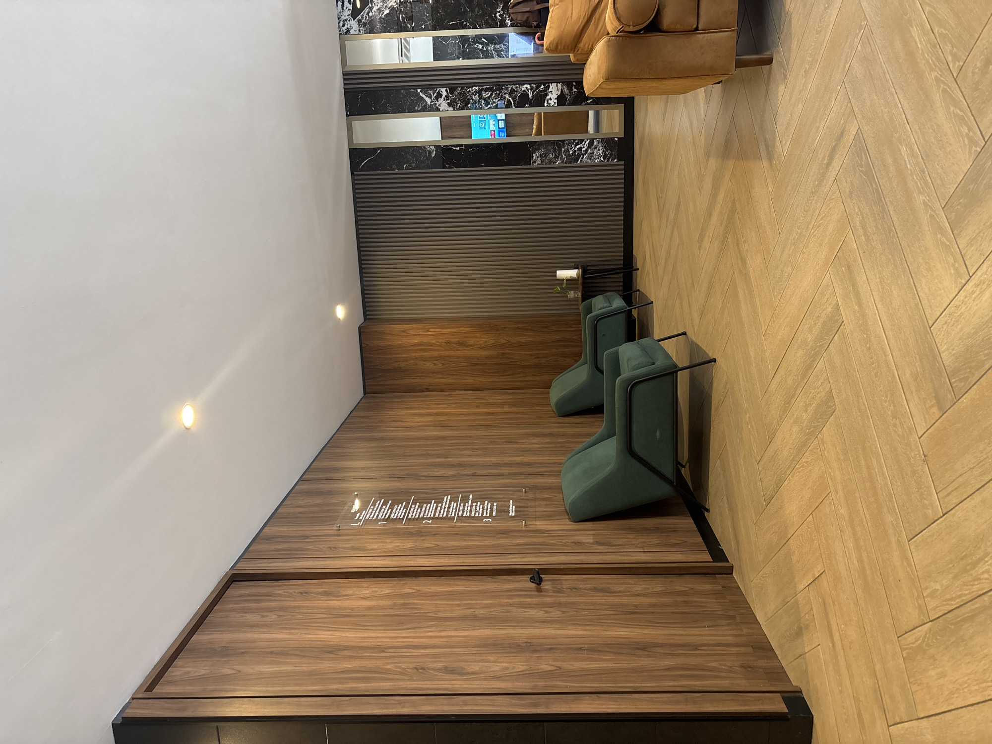 The image captures a portion of the interior of a business or hotel lounge in Boca del Río, Mexico. The design features a modern aesthetic with a blend of wood and dark stone-like surfaces. Two green chairs with black metal frames sit on a raised wooden platform, accompanied by small, clear side tables. A clear sign with text is affixed to the platform in front of the chairs. Above this area is a wall adorned with horizontal, textured panels and sections of black and white marbled material. Above these panels, a glimpse of a light brown leather sofa can be seen. The floor is tiled with light-colored wood in a herringbone pattern. The wall to the left is painted white. There are two recessed lights on the wall.