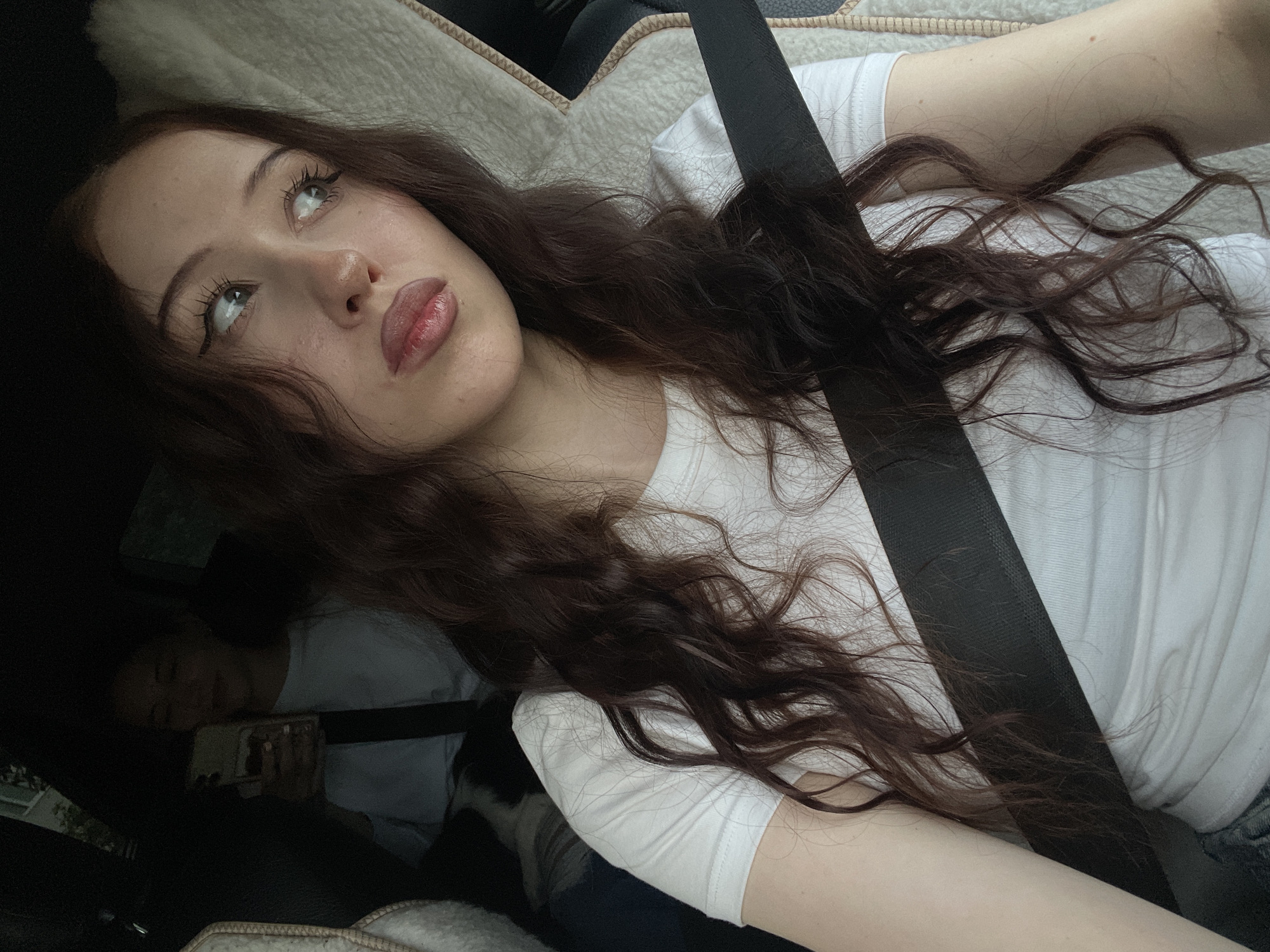The media file features a close-up, selfie-style shot of a young woman with long, wavy, dark brown hair. She is wearing a white t-shirt and a black seatbelt is diagonally across her chest. Her head is tilted back and her eyes are looking upwards, with a slight smile on her lips. The setting appears to be inside a vehicle, with the seatbelt and upholstery visible. The lighting suggests it is daytime. Another person can be seen in the background, partially obscured and appearing to be looking at a phone. The overall mood is relaxed and candid. There is no visible text in the image.
