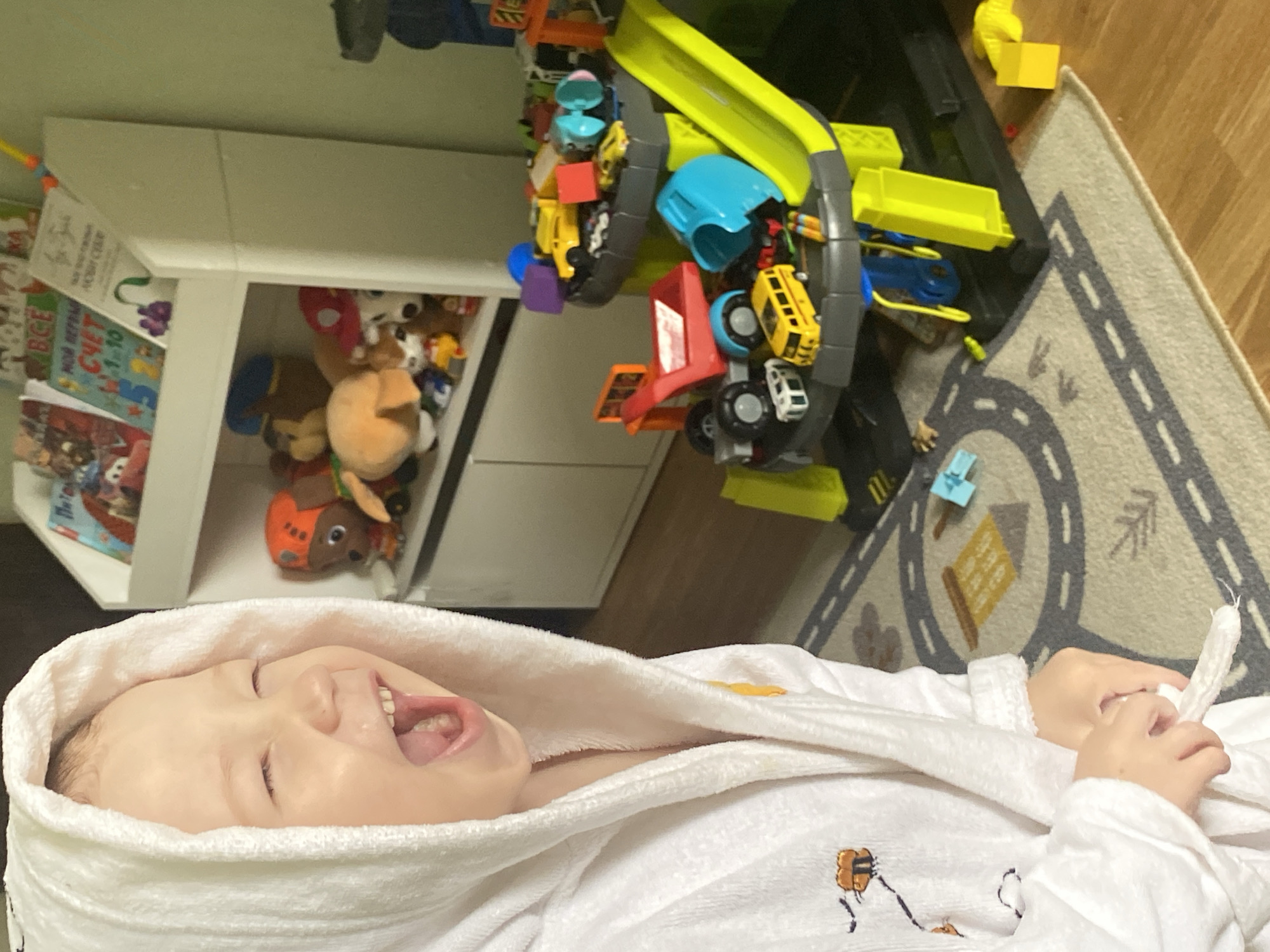A child wrapped in a white hooded robe is lying on a surface in what appears to be an indoor setting, potentially a home. The child is smiling and appears to be looking upwards. Above the child, there's a white shelf filled with toys, including stuffed animals and children's books. Beside the shelf, there's a toy car track with toy vehicles on it. A rug with road patterns is visible on the right side of the frame, adjacent to a wooden floor. There is no indication of the location being in Sochi, Russia.