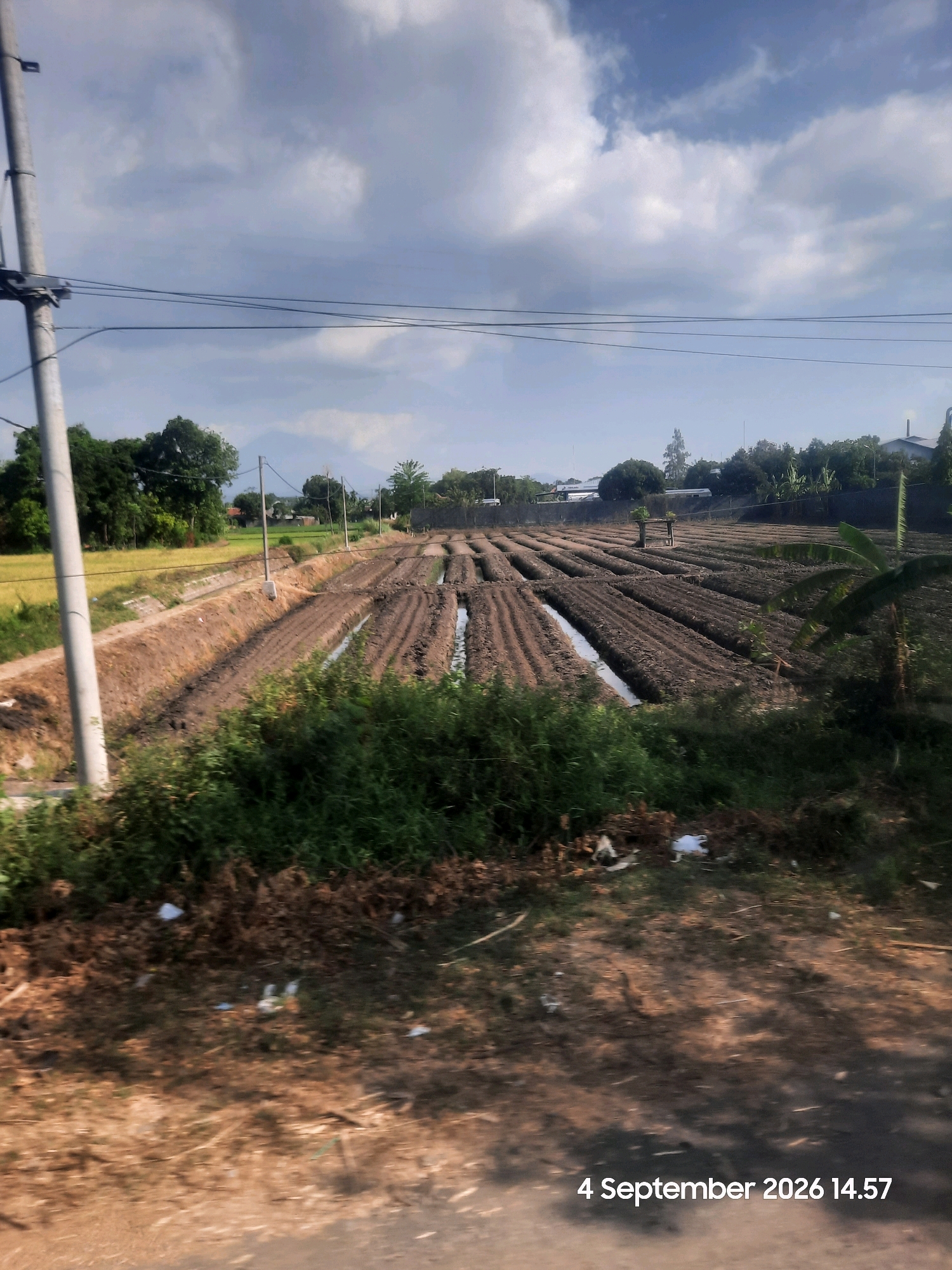 The image depicts a rural landscape in Bulang, Indonesia, characterized by agricultural fields and a cloudy sky. The foreground shows a dirt road with scattered debris, leading into a cultivated area with rows of raised soil beds, suggesting recent planting or preparation for crops. The fields appear to be irrigated, with water channels visible between the rows. In the background, lush green trees and vegetation flank the fields, and a distant mountain is faintly visible. A utility pole with electrical wires stands prominently on the left side of the frame. The overall scene conveys a sense of agricultural activity within a natural setting. The timestamp "4 September 2026 14.57" is superimposed on the lower right corner of the image.