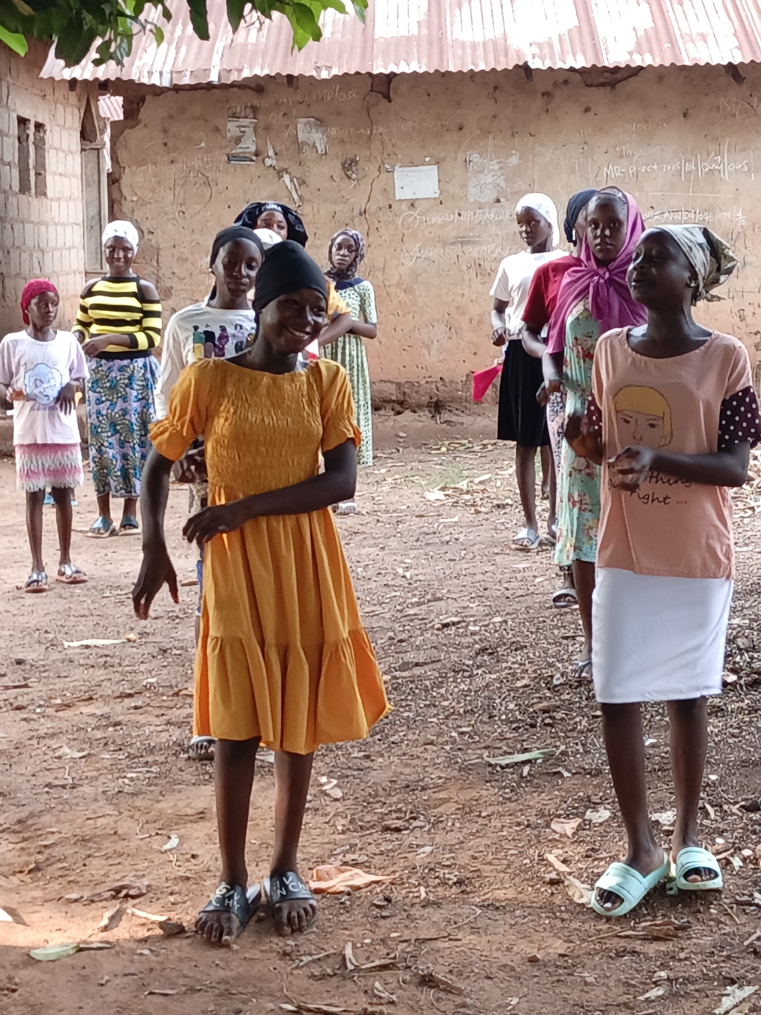 Akwanga, Nigeria, shows a group of approximately ten young women outdoors. Most of the women are wearing head coverings. One woman in the foreground is wearing a short, orange dress. Another woman in the foreground is wearing a pink t-shirt and white skirt. The background includes a building with a corrugated metal roof and an unpaved, dirt ground.