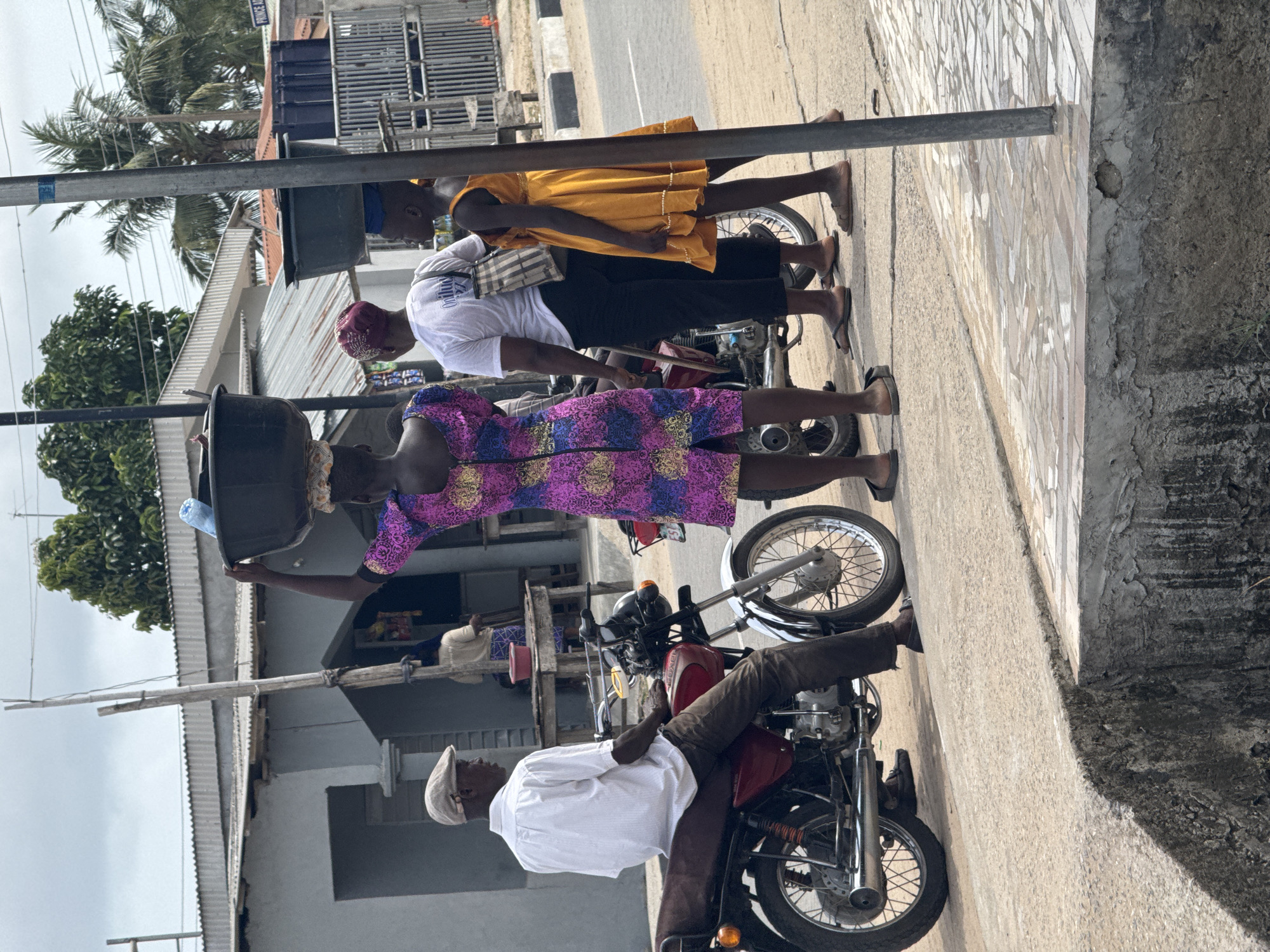 The image depicts a street scene in Badagry, Nigeria. Three women stand on a sidewalk next to two motorcycles parked along the curb. One woman is carrying a large black bucket on her head. She wears a colorful patterned dress. Another woman wears a white shirt, black pants, and a red head covering. The third woman wears a yellow dress. On one of the motorcycles, a man sits. He wears a white shirt, brown pants, and a white cap. Buildings line the opposite side of the street, and trees are visible in the background. A utility pole is near the building side of the street.