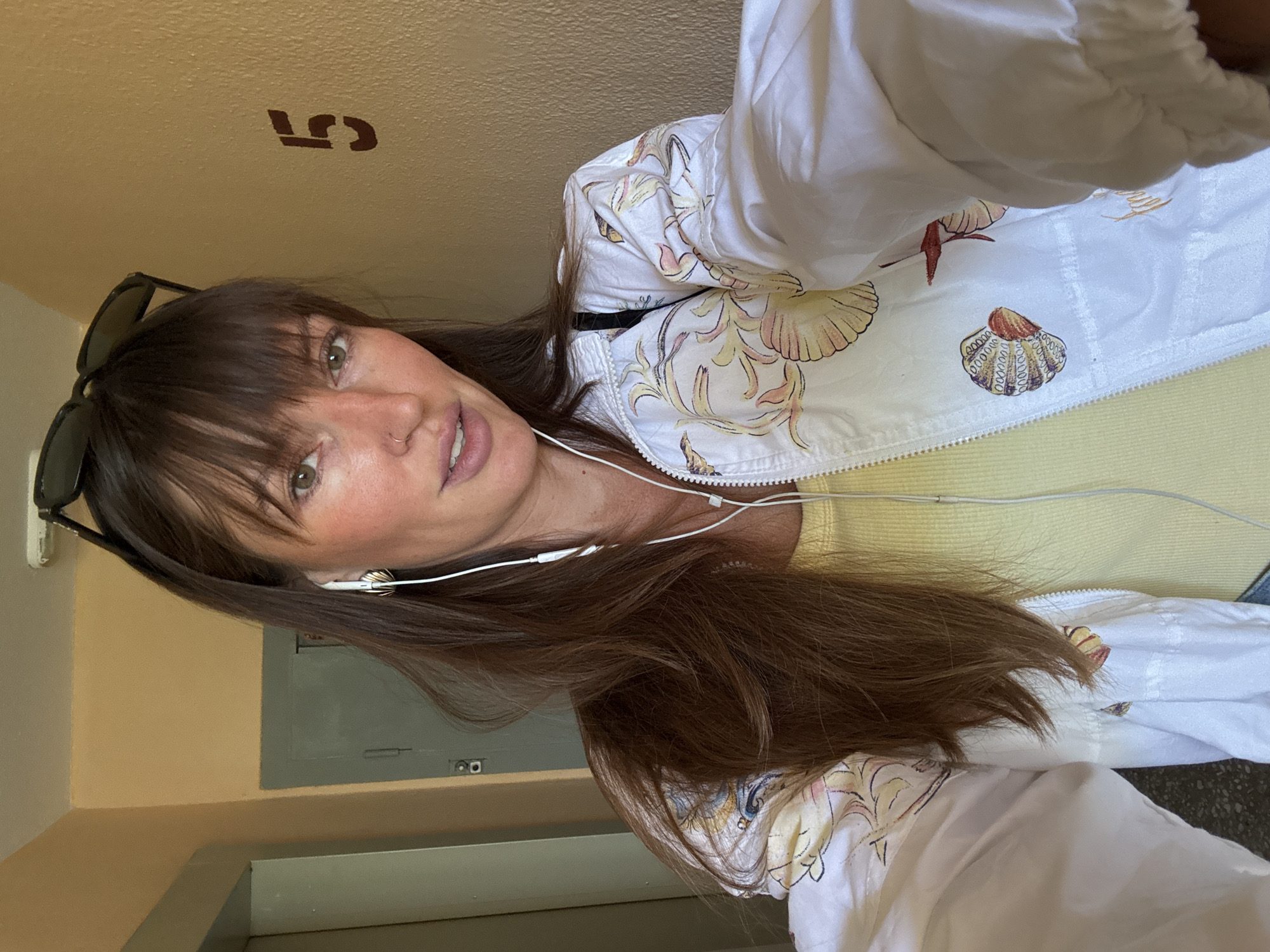 A woman is taking a selfie in a hallway with beige walls and a gray door. She has long brown hair, bangs, and is wearing sunglasses on top of her head, white earbuds, and a white jacket with a floral pattern over a yellow shirt. A dark brown number "5" is stenciled on the wall behind her. The overall impression is that she is in a residential building.