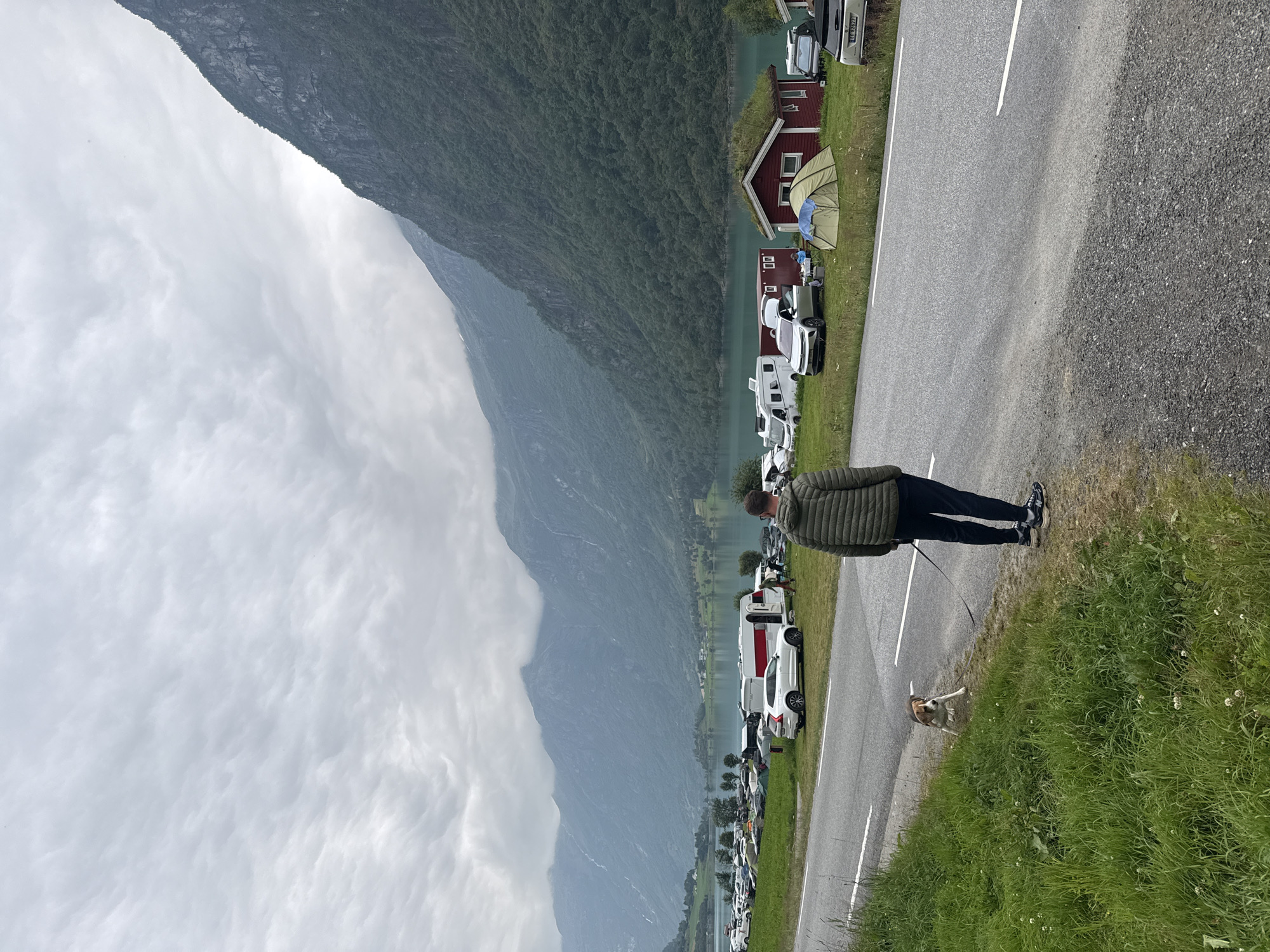 A man wearing a green puffer jacket and dark pants stands on the right side of a two-lane asphalt road, facing away from the camera and looking towards a body of water and mountains. A small, tri-colored beagle on a leash stands to his left, closer to the grassy shoulder. Across the road, there is a campground situated by a lake, with numerous tents, campers, and cars. A red building with a grass roof is visible in the campground, along with a green tent. The background features a vast, calm body of water and a series of imposing, tree-covered mountains under a cloudy sky, indicative of the scenic landscape in Olden, Norway. The scene appears to be a typical summer day in a popular tourist area.