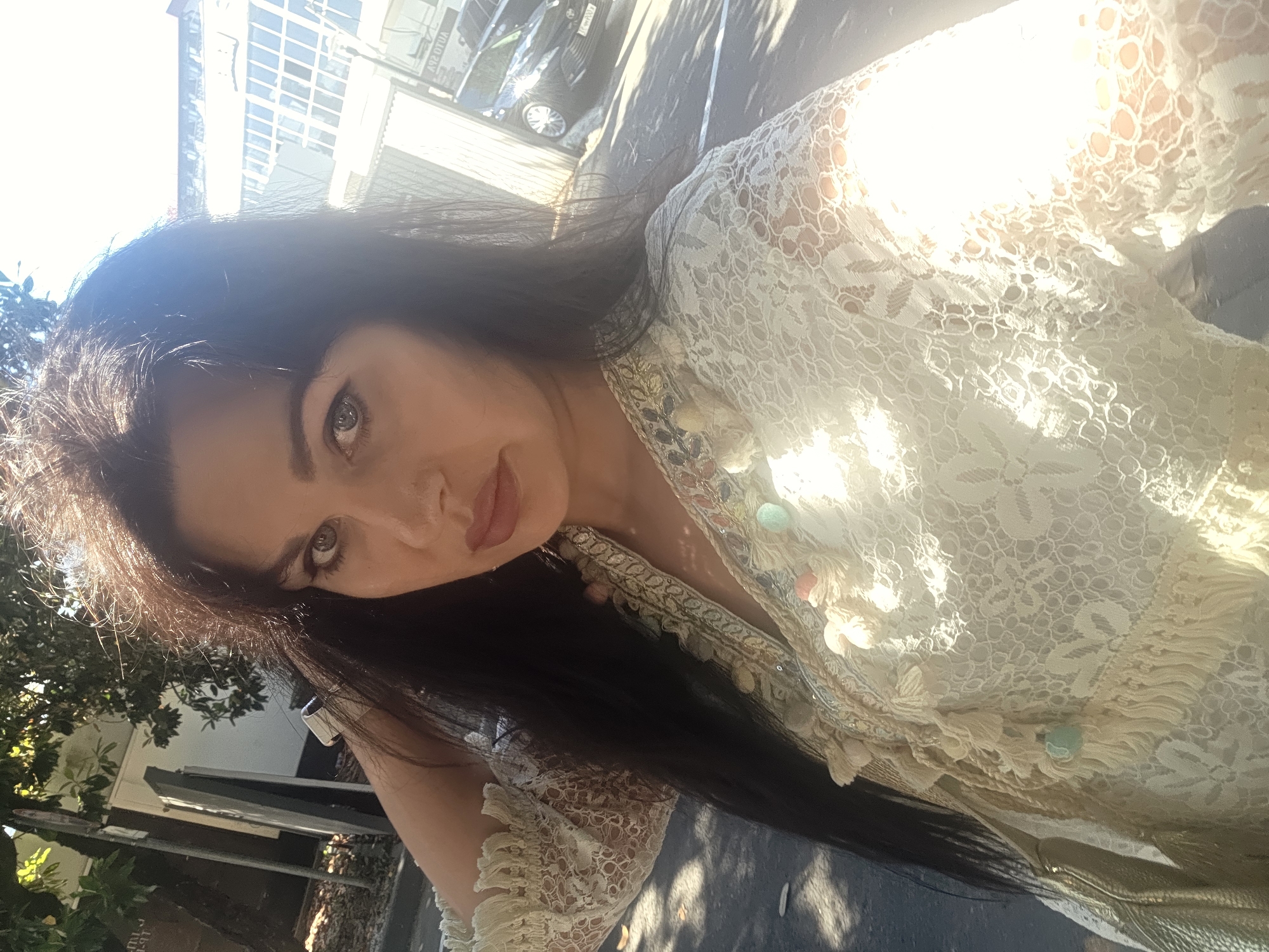 A woman with long, dark, wavy hair and light-colored eyes is looking into the camera. She is wearing a light-colored lace top with intricate embroidery and embellishments. Her left arm is bent, and her hand is near her head. The background shows a sunny outdoor scene with trees, a building with large windows, and a car. The presence of "AUTO SPA" signage suggests a commercial area. The lighting indicates it is daytime. No specific landmarks of Sochi, Russia are identifiable.