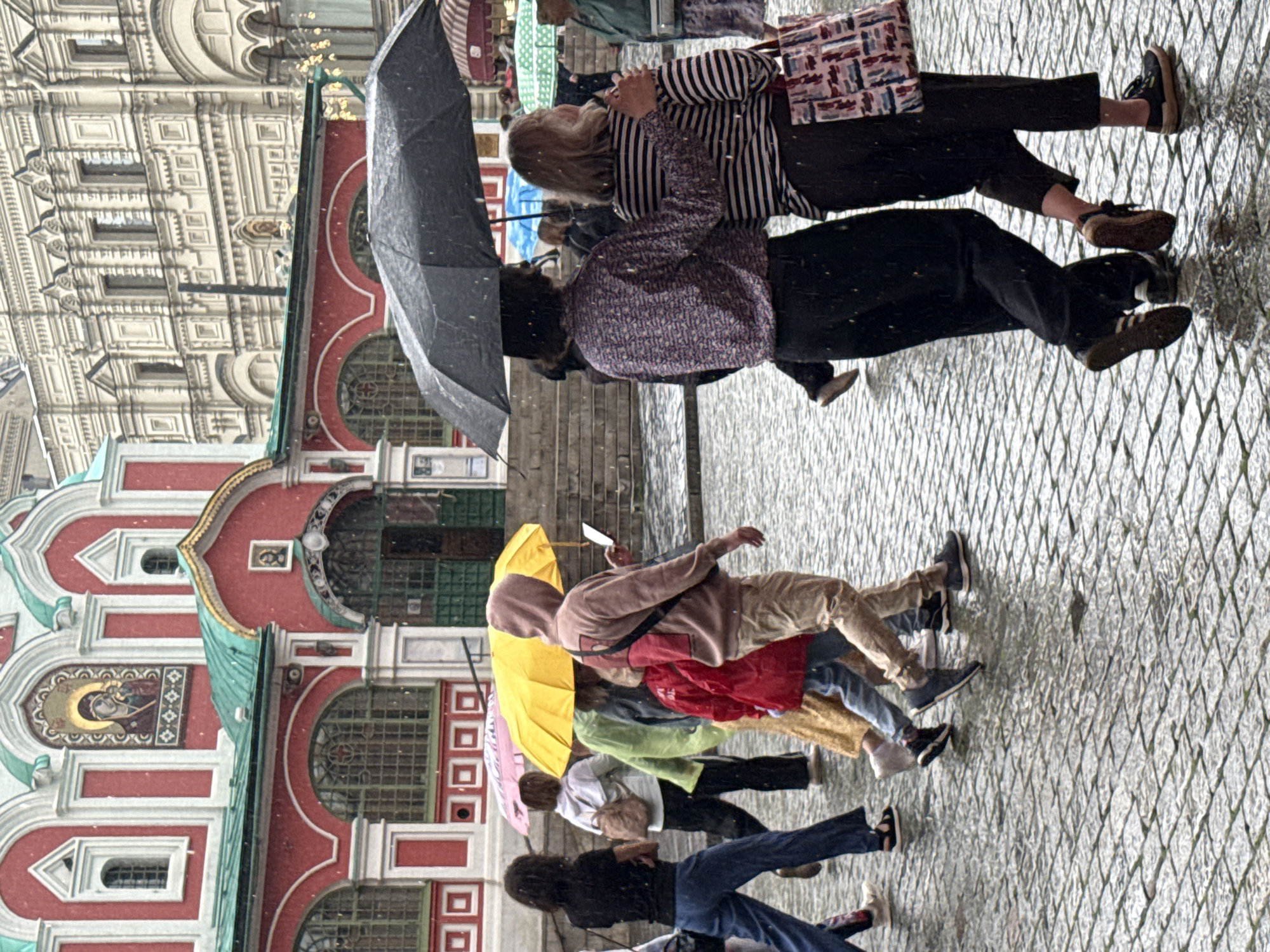 The image depicts a rainy day scene on Red Square in Moscow, Russia. Several individuals are walking across the cobblestone ground, many of them holding umbrellas. In the background, the colorful facade of Saint Basil's Cathedral is visible, along with elements of the historical buildings that surround the square. The weather is overcast, and raindrops are evident, creating reflections on the wet pavement. The people are dressed in casual attire, and some are carrying bags. The overall atmosphere suggests a typical tourist experience during inclement weather in a prominent Moscow location.