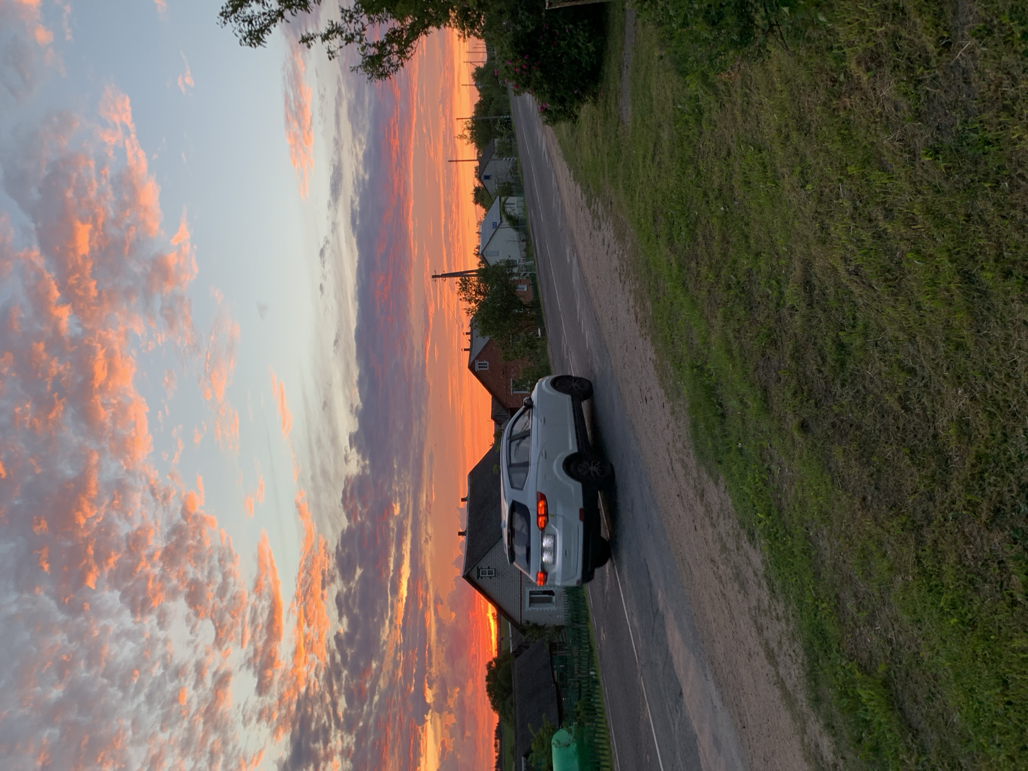 The image depicts an outdoor scene at sunset, located in the city of Відзаўскі сельскі Савет, Belarus. The upper two-thirds of the frame are dominated by a vibrant sky, displaying intense orange and red hues concentrated towards the horizon on the left, transitioning to lighter pinks, blues, and grays at the top right. Scattered, textured clouds are illuminated by the low sun.
Below the horizon line, a rural or semi-rural landscape unfolds. Multiple single-story detached houses with various roof and facade colors are visible, interspersed with trees and green foliage. A prominent grey-sided house with a darker roof stands to the left of the frame, near a green cylindrical tank and a wooden fence.
A white crossover SUV, with illuminated red taillights and white reverse lights, is positioned on a paved road that traverses the midground. The vehicle is angled slightly away from the viewer, traveling towards the left side of the frame. The road appears to be a two-lane asphalt surface, somewhat worn, with dirt shoulders on both sides.
In the foreground, to the left of the road, a narrow strip of grass and dirt is visible. To the right of the road, a wider, uneven grassy embankment extends into the lower right corner, featuring a dark green bush with small pink flowers. No people are visible in the scene.