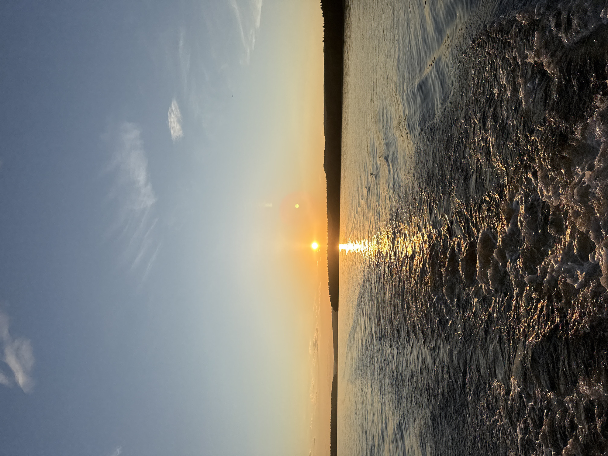 The image depicts a sunset over a wide body of water, likely the Votkinsk Reservoir near Votkinsk, Russia. The perspective is from the stern of a moving vessel, evidenced by a prominent, turbulent wake extending from the foreground towards the distant horizon. The sun, a bright golden-orange orb, is positioned low in the sky slightly to the right of center, casting a strong, shimmering reflection across the water, predominantly on the right side of the wake. A lens flare is visible as a distinct bright spot to the upper left of the sun. The sky transitions from a deep blue at the top to a lighter, warm yellow-orange hue near the horizon, with sparse, wispy white clouds scattered throughout. A dark, forested landmass forms a silhouette against the vibrant sky along the horizon, with a noticeable dip in the distant landform appearing in the center. The water in the foreground, particularly within the wake, exhibits significant agitation and frothy whitecaps, which gradually diminish further from the viewer. No individuals, identifiable subjects, or specific man-made objects other than the implied vessel are present in the frame. The overall environment conveys a tranquil evening setting.