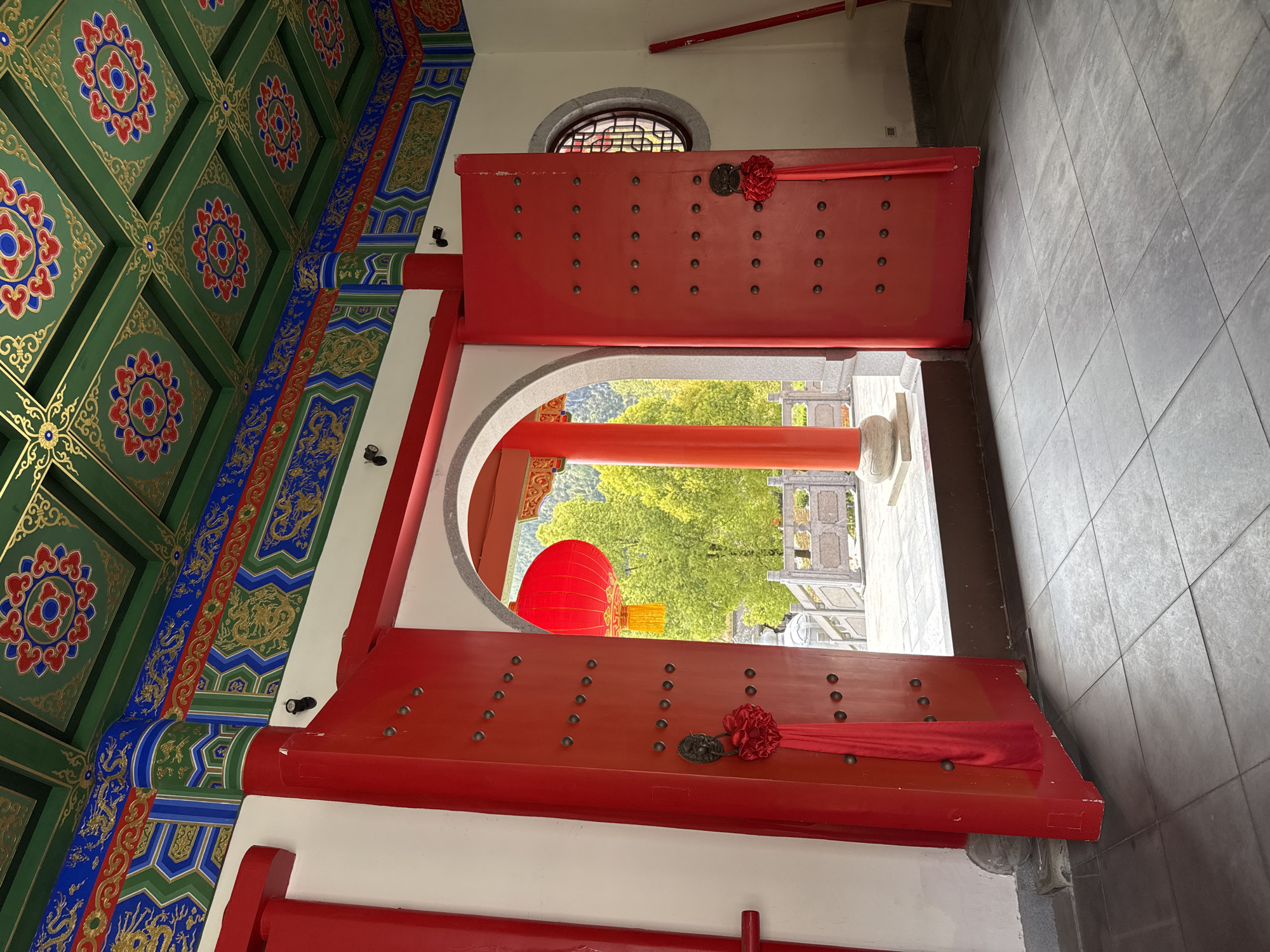 View through a red door with ornate detailing on the ceiling and door. Outside the door are trees and building in Qianjiang, China.