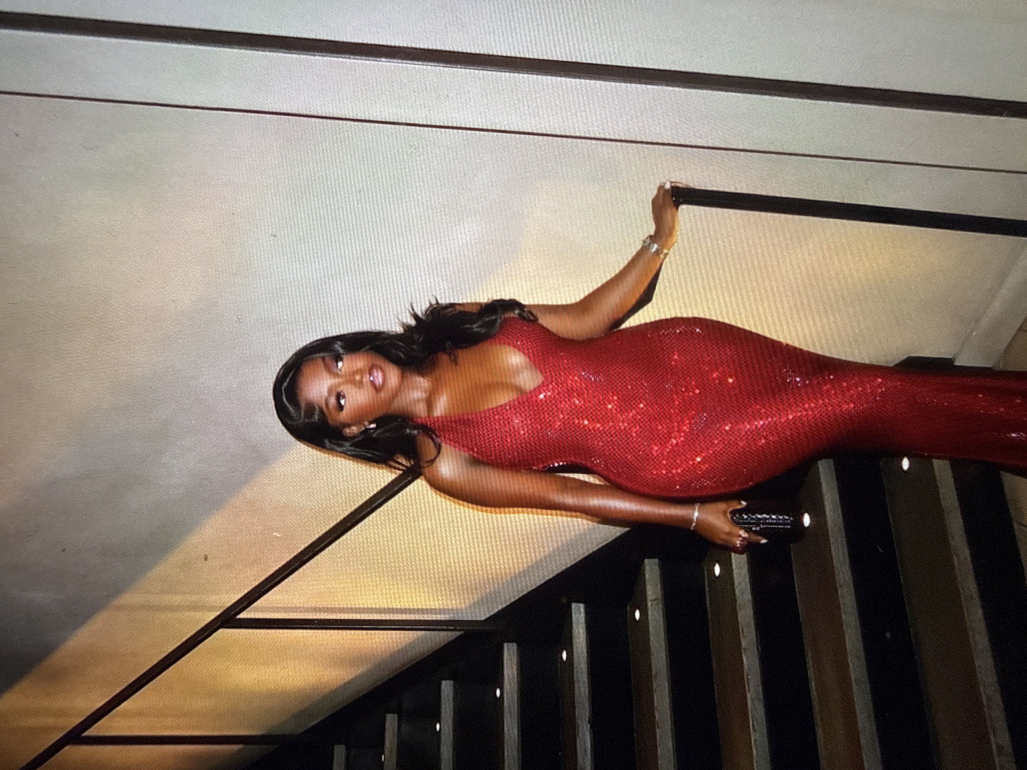 A Black woman wearing a sparkling red halter dress stands on a staircase, holding onto a railing. The woman has long, dark hair and is wearing jewelry on her wrist and ears. She is holding a black clutch in her left hand. The staircase is dark wood with metal railings. The background appears to be a wall with a textured surface, and there is lighting from above casting shadows on the wall and staircase. The scene suggests an indoor or possibly outdoor event at night. There is no information in the image to indicate the location is the city of Abuja, Nigeria.