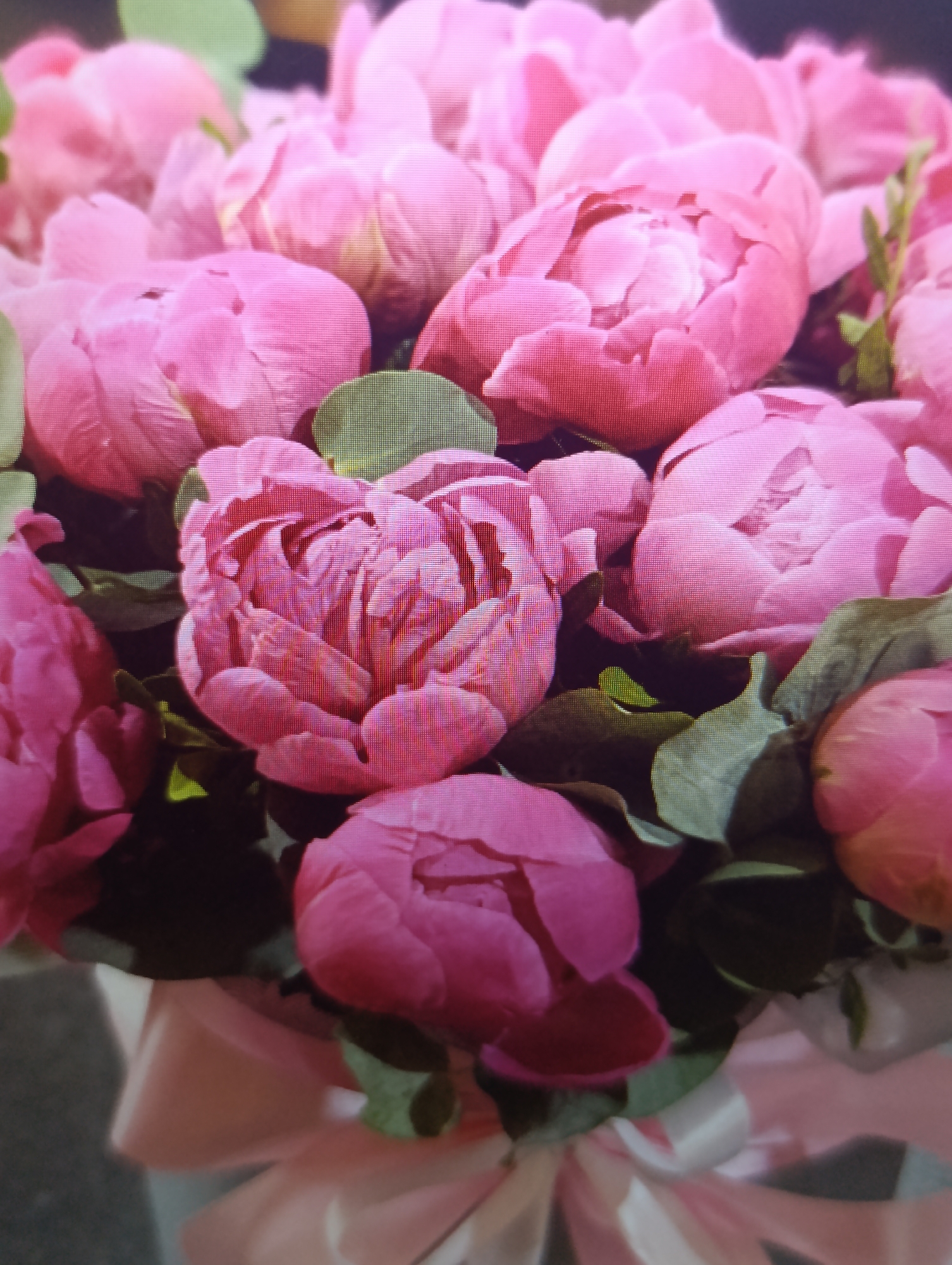 A close-up, slightly blurry shot captures a bouquet of vibrant pink peonies. The flowers are densely packed, showcasing their ruffled petals in various stages of bloom. Lush green leaves peek through the blossoms, adding contrast to the soft pink hues. The bottom of the image reveals hints of a pink ribbon and possibly a box or vase, suggesting the bouquet is arranged for gifting or display. The background is a dark, indistinct blur. The scene is indoors, with artificial lighting creating soft shadows and highlighting the textures of the flowers.