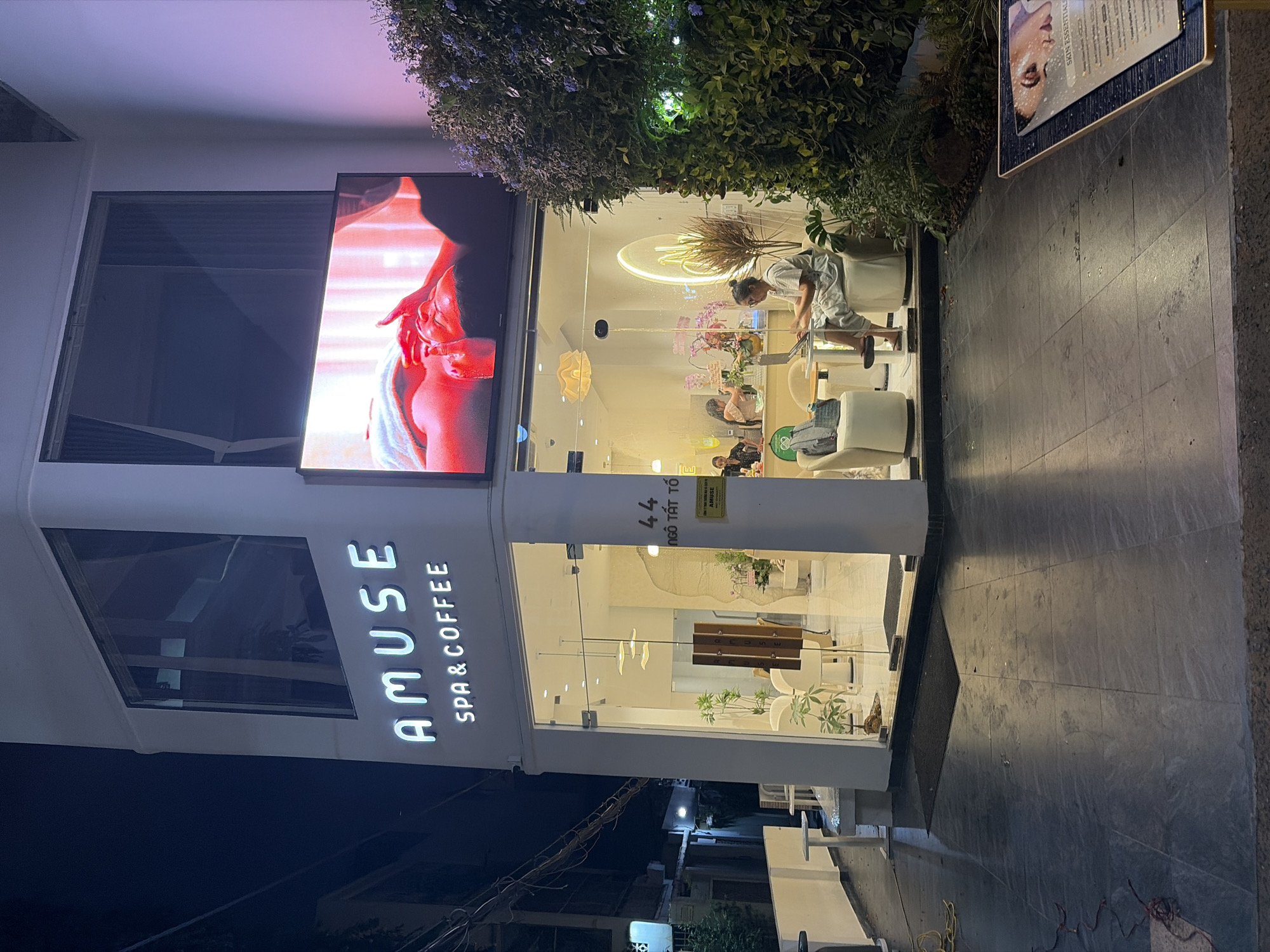 This image presents the nighttime exterior of "AMUSE SPA & COFFEE," a commercial establishment identified by its illuminated signage. The building features a modern, white facade with large glass windows on the ground floor, revealing a brightly lit interior. The main sign displays "AMUSE" in large capital letters, with "SPA & COFFEE" centered directly below. To the right of this signage, a large digital display screen exhibits a close-up image of a person's face receiving a spa treatment, illuminated with a reddish-orange hue.
Through the glass windows, the interior shows a contemporary, minimalist design with light-colored walls and furniture. A woman is seated at a small round table on the right side, actively using a laptop. She is wearing light-colored, possibly striped, casual attire. Two additional individuals are partially visible further inside near a service counter. The interior is furnished with white, upholstered chairs, small round tables, potted plants, and modern decorative ceiling lights. A large, glowing stylized "M" from the Amuse branding is visible on an interior wall. An address plaque near the entrance displays "44 Ngô Tất Tố."
The exterior ground area is paved with dark gray tiles. Two black doormats are placed at the entrance. To the left of the building, a low white wall extends with a small bench and a white table. Further left, a dimly lit street or alleyway is visible in the background, with overhead wires. To the right of the building, a dense vertical garden or large bush covered in small purple flowers borders the structure. In the immediate foreground on the right, a freestanding display stand with "SKIN ESSENTIALS" text and an image of a face is partially visible. Loose yellow and red/purple wires are present on the pavement in the left foreground. The location is specified as Thủ Đức, Vietnam, with the visible street address being 44 Ngô Tất Tố.