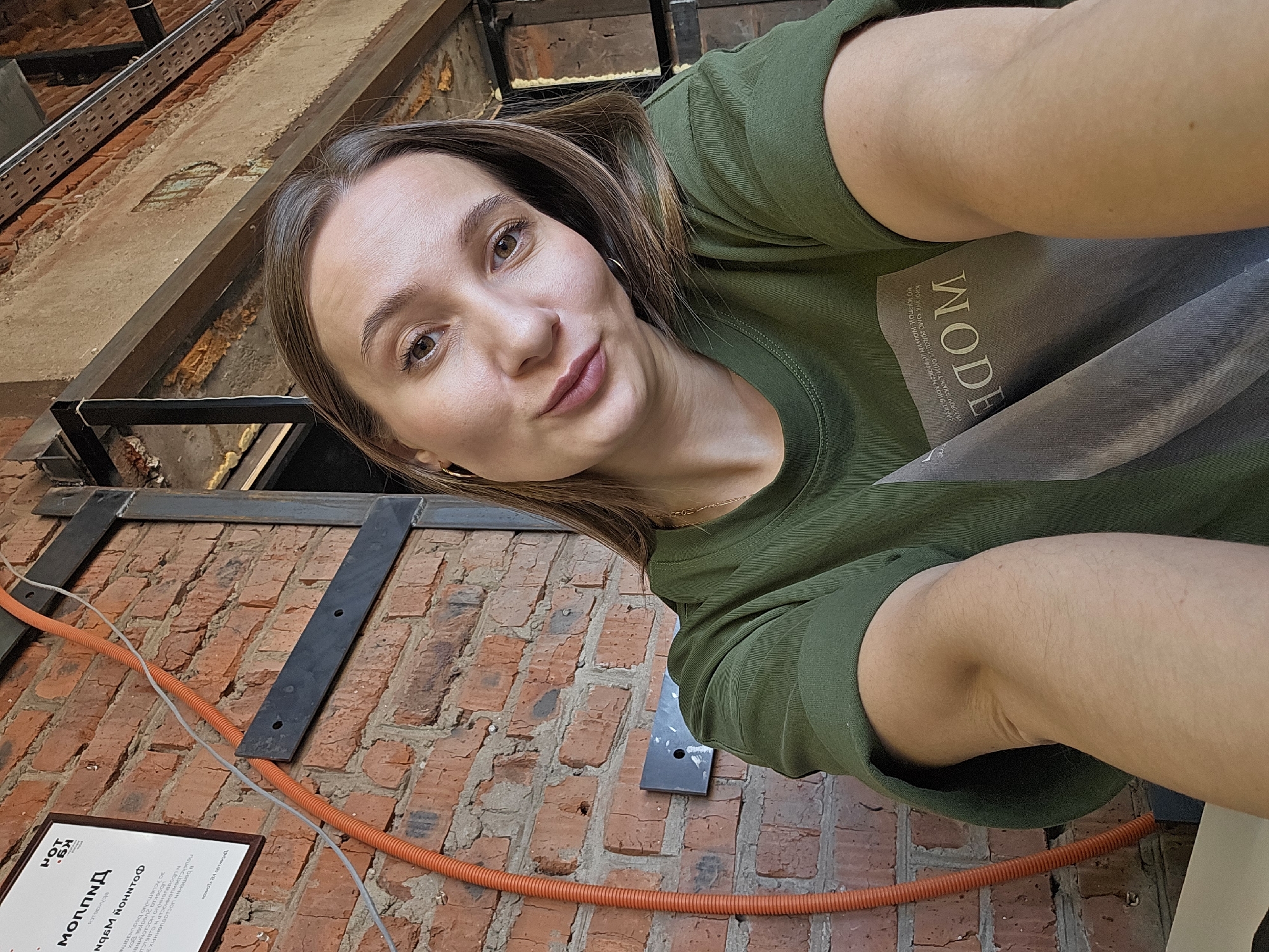 A woman with shoulder-length brown hair and fair skin takes a selfie at an unusual angle. She wears a green shirt with the word "WODE" on it. Behind her, there is a brick wall with metal beams and orange conduit running across it. A sign is partially visible in the lower left corner. Based on the text on the sign, this image was taken in Chaikovsky, Russia.