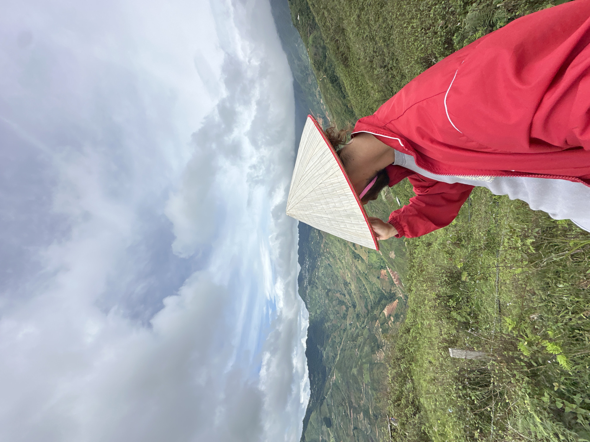 A person wearing a conical hat and a red jacket stands on a grassy hillside, obscuring their face with the hat. The background features lush green mountains with terraced fields, indicative of a rural mountainous region. The sky is cloudy and overcast, suggesting a possibly humid or rainy day. This scenery is characteristic of Ta Xua, Vietnam, known for its breathtaking landscapes and mountainous terrain.