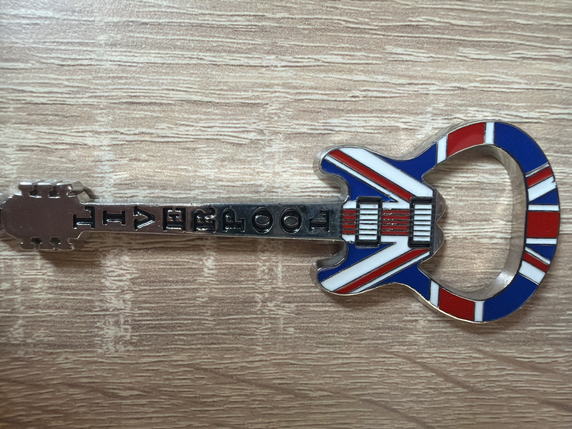 An overhead shot displays a metallic, guitar-shaped souvenir item resting on a light brown wooden surface. The item's body is intricately designed with the pattern of the Union Jack flag, featuring bold red crosses and white diagonal lines against a blue background, all outlined in silver. The flag design is enamel-filled, providing distinct coloration.
The neck of the guitar-shaped object is silver and features raised, block capital letters that spell out "LIVERPOOL". The headstock end of the guitar is also silver. The item's shape and design suggest it could be a keyring, bottle opener, or a decorative magnet, commemorating the city of Liverpool, United Kingdom, which is famous for its musical heritage, particularly The Beatles.
The wooden surface beneath has a fine, horizontal grain pattern. The lighting is soft and even, highlighting the metallic sheen and colorful enamel of the object. The image was captured in Plovdiv, Bulgaria.