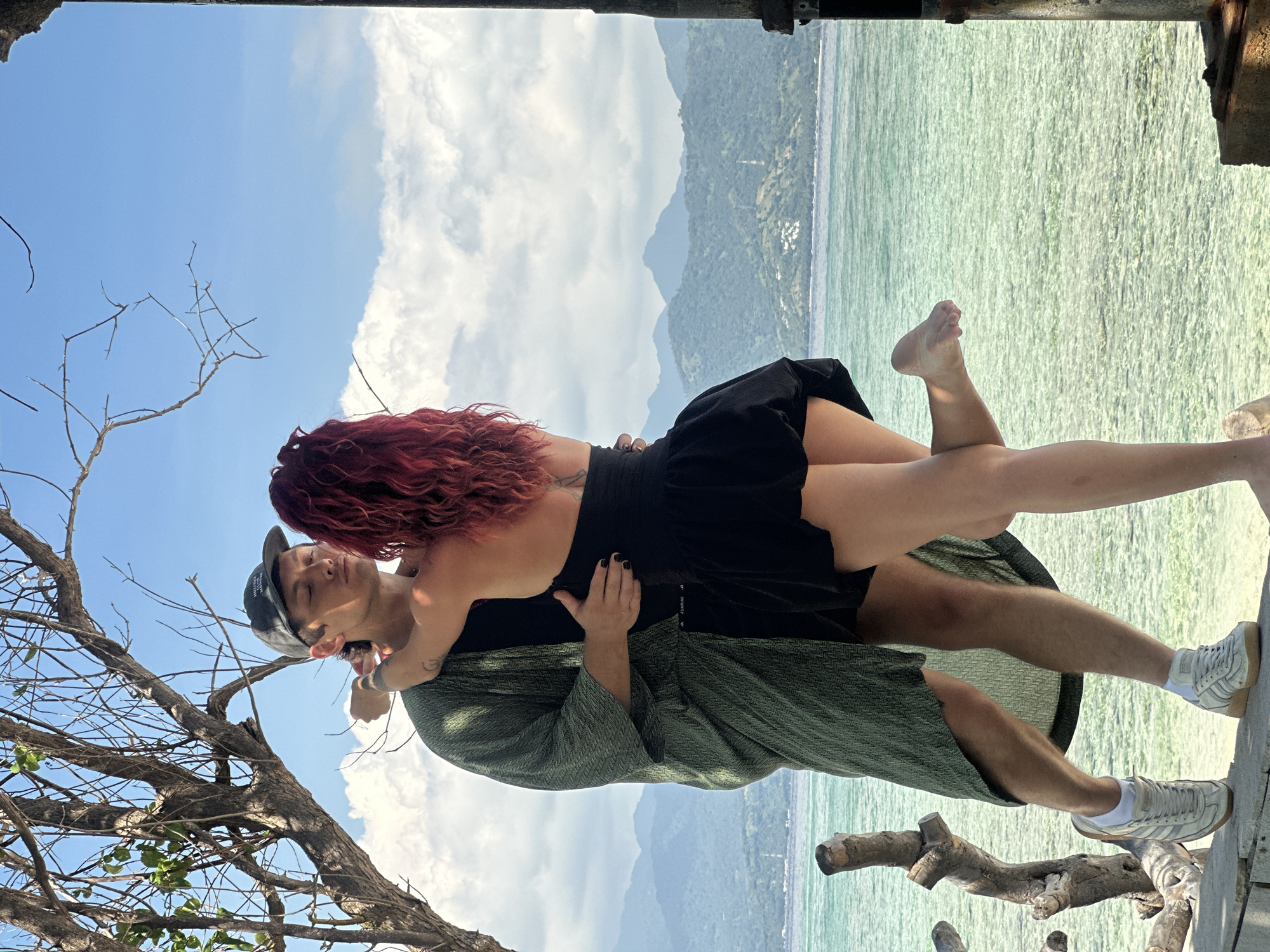 A couple embraces intimately on a wooden structure in Unknown, Indonesia. The woman, with vibrant red curly hair, wears a black dress. The man, sporting a cap and green patterned outer garment, holds her. The background features a light turquoise sea, a mountainous landscape, and a partly cloudy blue sky. A bare tree branch is visible on the left.