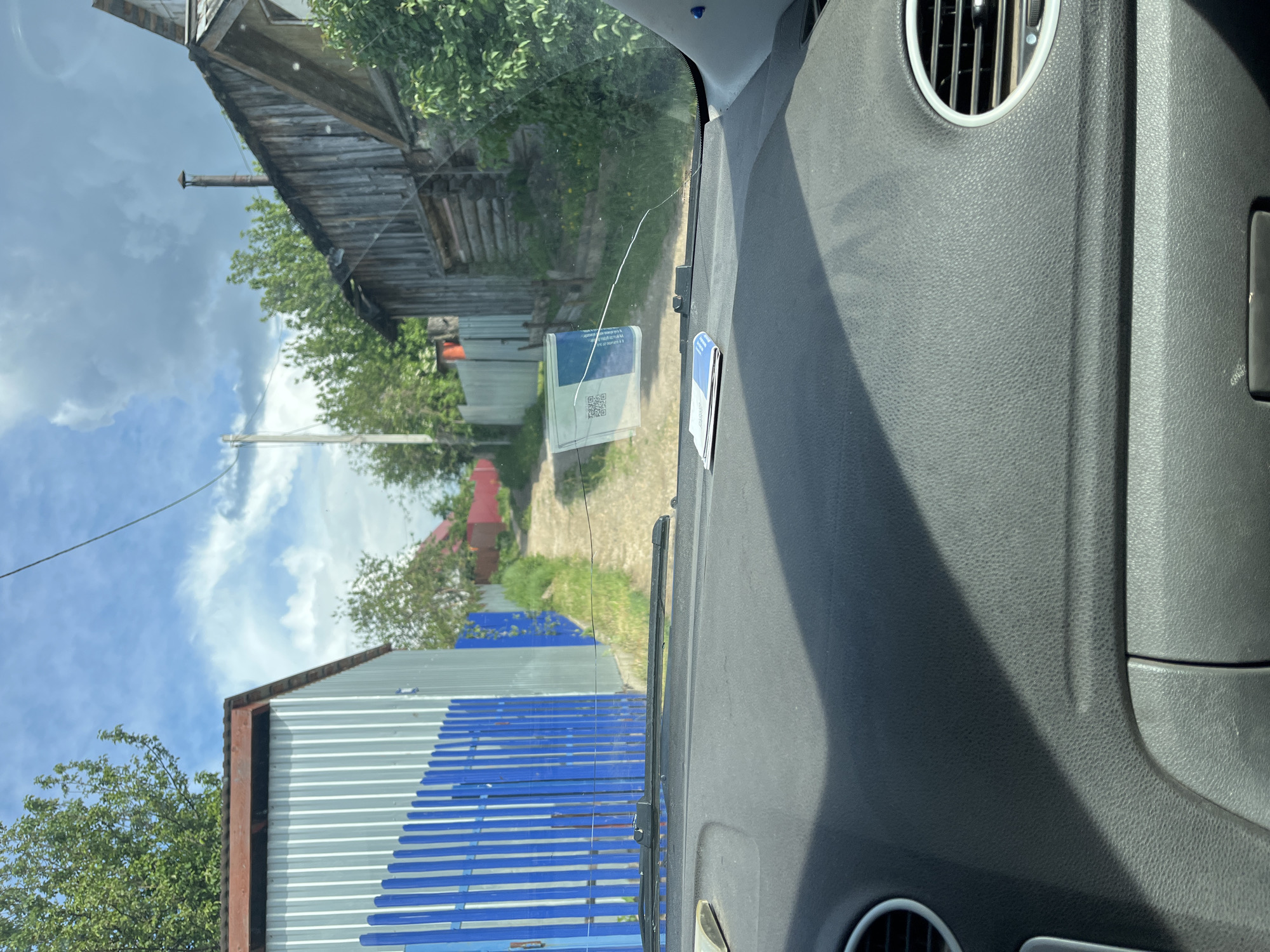 The image is taken from inside a vehicle, looking out through the windshield towards a rural scene in Chaikovsky, Russia. The dashboard of the vehicle is visible, featuring vents and a gray textured surface. Outside, a dirt road leads into the distance, flanked by buildings and vegetation. On the left, a building with blue and gray corrugated metal siding is visible. Further down the road, there's an older wooden building and glimpses of other structures with red roofs. The sky is partly cloudy. A crack is present in the windshield, along with a sticker.