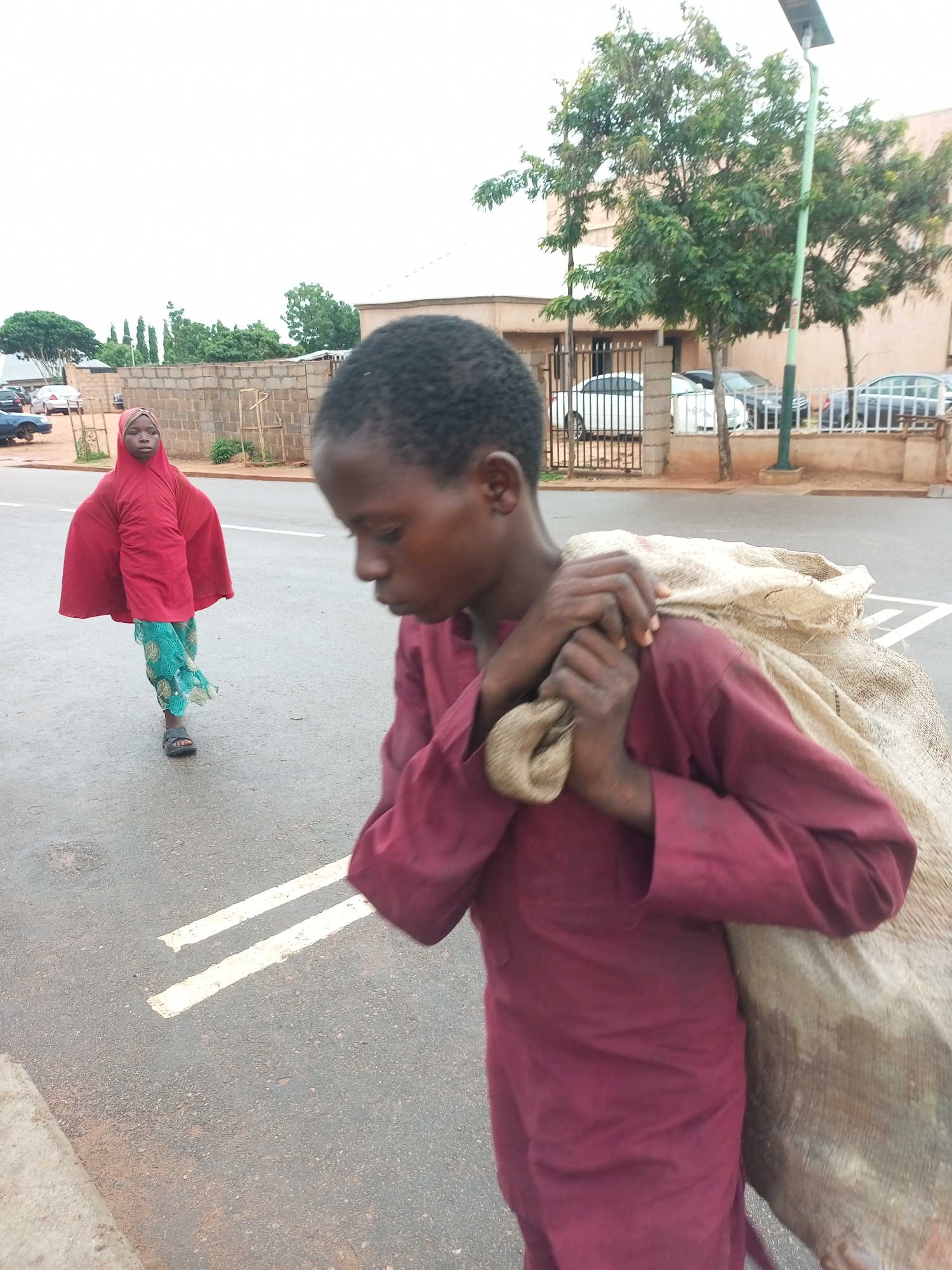 In Gombe, Nigeria, two children walk along a paved road with white lane markings. The child in the foreground, a boy, walks towards the right side of the frame, facing away from the viewer. He is carrying a large, light-colored sack over his right shoulder and appears to be wearing a maroon long-sleeved garment. His hands are grasping the sack. Behind him and to his left, a girl walks towards the left side of the frame, facing the viewer. She is wearing a bright red hooded garment and a patterned green skirt. She has dark hair and is looking towards the camera. The road is wet, suggesting recent rain. In the background, there is a low brick wall, some trees, buildings, and parked cars. A lamppost with a solar panel is visible on the right side of the frame. The sky is overcast.