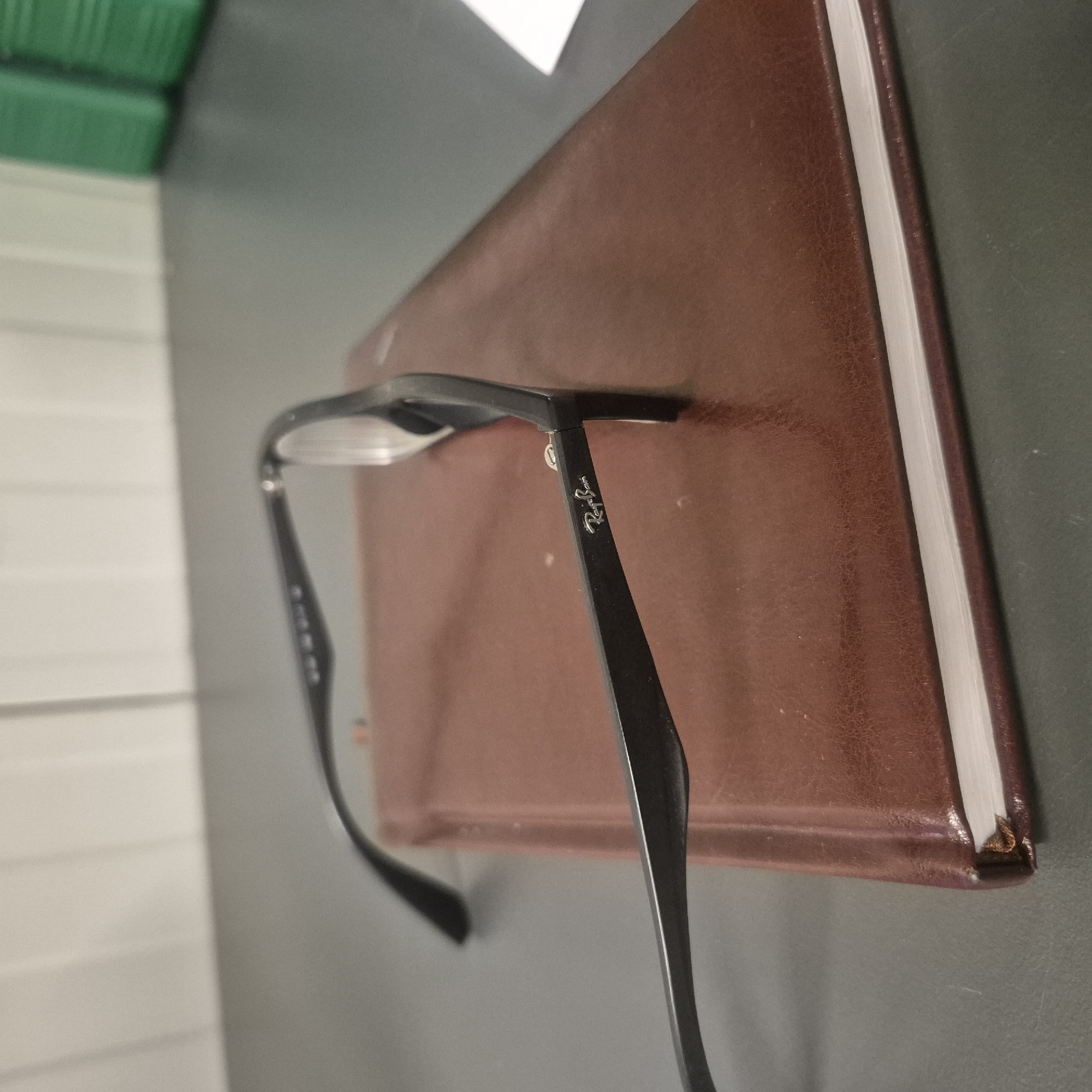 In Nowa Wieś Książęca, Poland, on a dark gray desk, a pair of black-rimmed eyeglasses with clear lenses rests on top of a brown, faux-leather bound notebook. The notebook's pages are visible, and the binding shows slight wear at the corner. The eyeglasses are positioned diagonally across the notebook, with the left arm extending towards the viewer. The brand "Ray-Ban" is subtly embossed in silver on the side of the left arm. In the background, a light-colored wall with vertical slats is visible, and to the right, two green plastic containers are present. The scene appears to be indoors, possibly in an office or study.