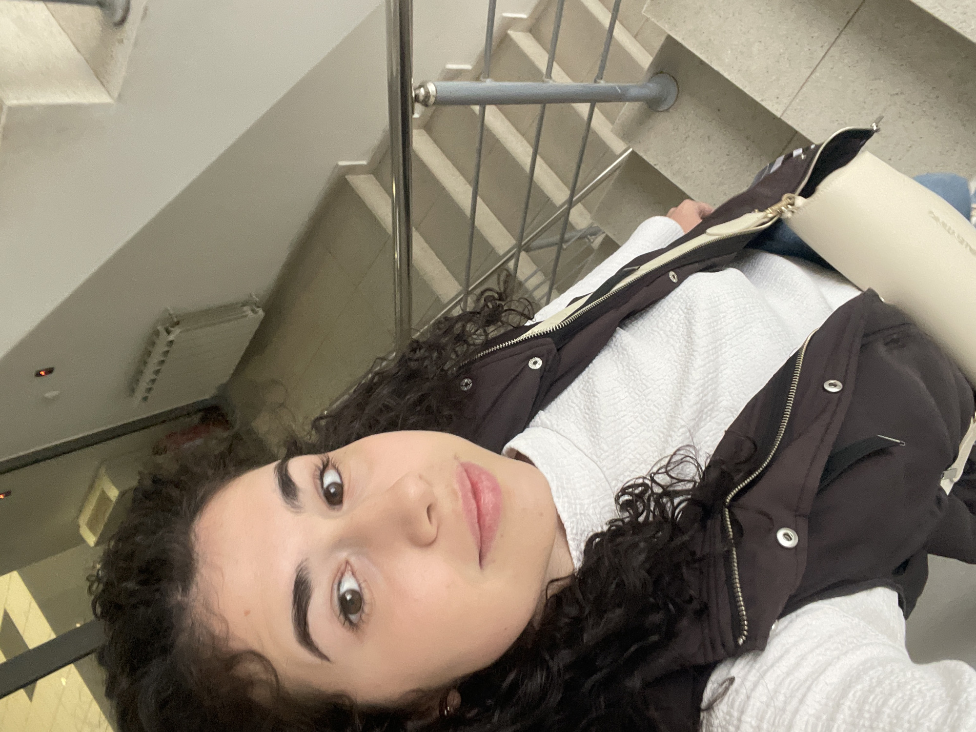 An indoor selfie captures a young woman with dark, curly hair, light brown eyes, and prominent eyebrows, looking directly at the camera. She wears a white textured long-sleeved top and a dark brown or black puffer vest with a prominent zipper and silver snaps. Her lips have a soft pink tint.
The woman is positioned on or near a staircase, which serves as the background. The staircase features grey concrete steps and a metal railing with vertical bars, casting distinct diagonal shadows onto the steps. A light beige or off-white handbag with a gold-colored clasp is visible next to her, with faintly discernible text, possibly a brand name, on its surface.
The setting appears to be a modern building interior, likely a stairwell or landing, characterized by plain white walls and a ceiling. A small, wall-mounted unit, possibly a radiator, is visible in the upper background. The lighting is soft and even, suggesting indoor ambient light. The woman's expression is neutral and calm. The location context for this image is Sofia, Bulgaria.