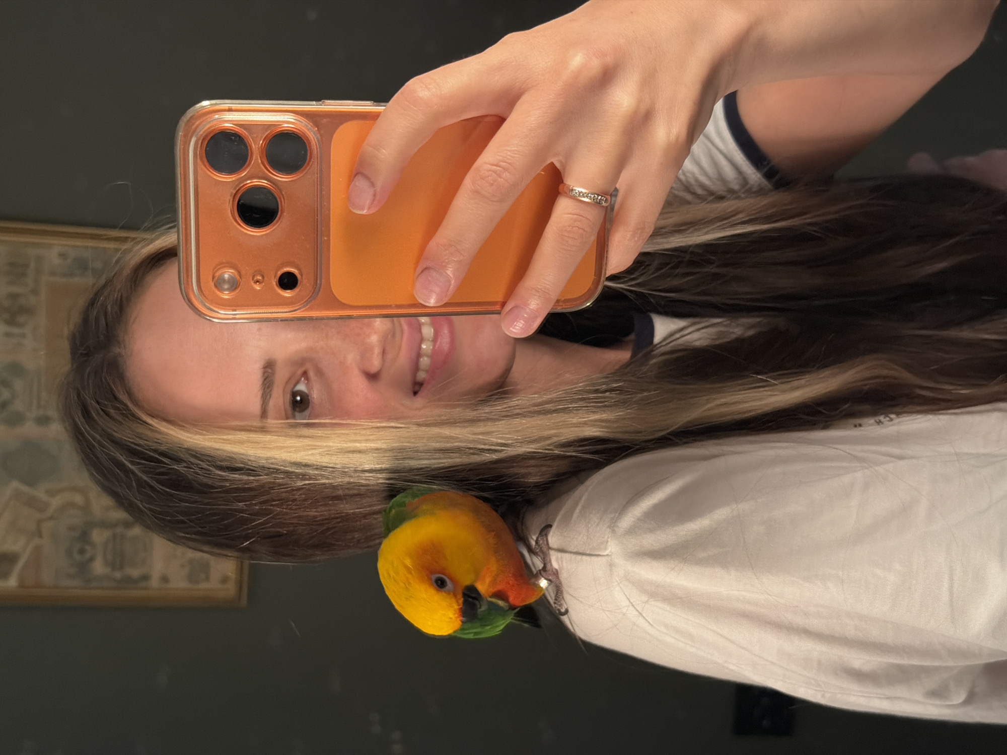 A young woman with long, blonde-streaked brown hair takes a selfie in a room. She is smiling at the camera, which she holds in her right hand. She is wearing a white t-shirt with a navy blue trim around the collar. A small, brightly colored parrot is perched on her left shoulder. The parrot has yellow, orange, and green feathers. The woman is wearing a ring on her left ring finger. In the background, a framed picture hangs on a dark-colored wall. The image does not provide any specific details about the city of Khimki, Russia.