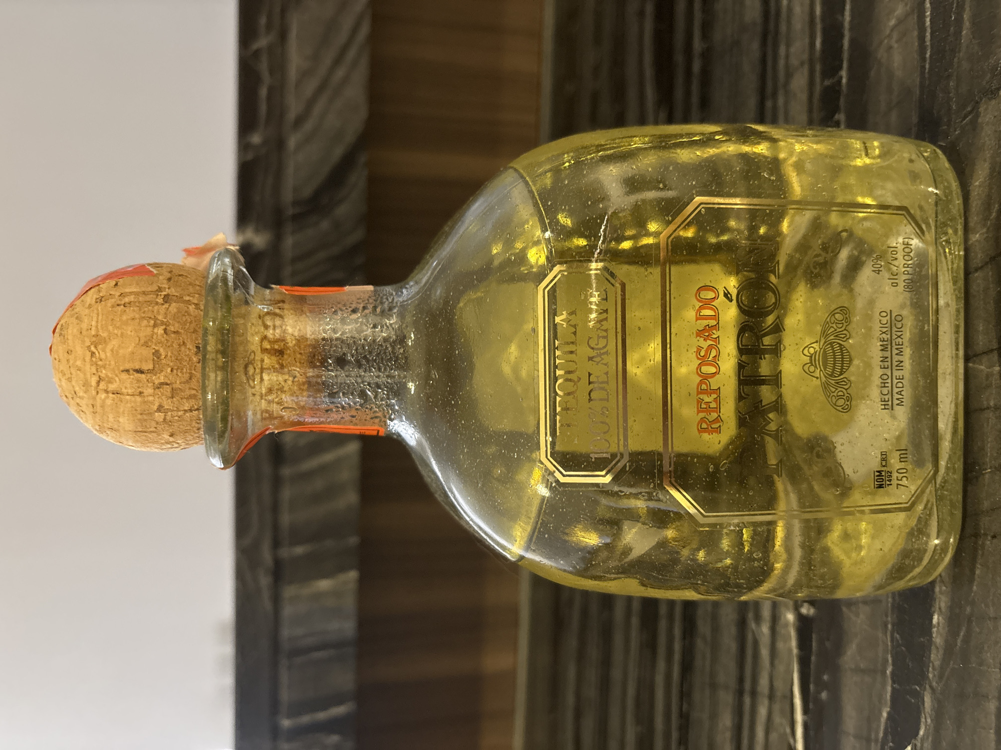 A clear glass bottle of Patrón Reposado tequila sits on a dark, reflective surface. The bottle has a distinctive shape with a cork stopper. A label on the bottle reads "TEQUILA 100% DE AGAVE REPOSADO PATRON". The liquid inside is a pale golden color. The background consists of a blurred wooden surface and a lighter wall. The scene is indoors, and there is no indication of the city of Dubai or the United Arab Emirates.