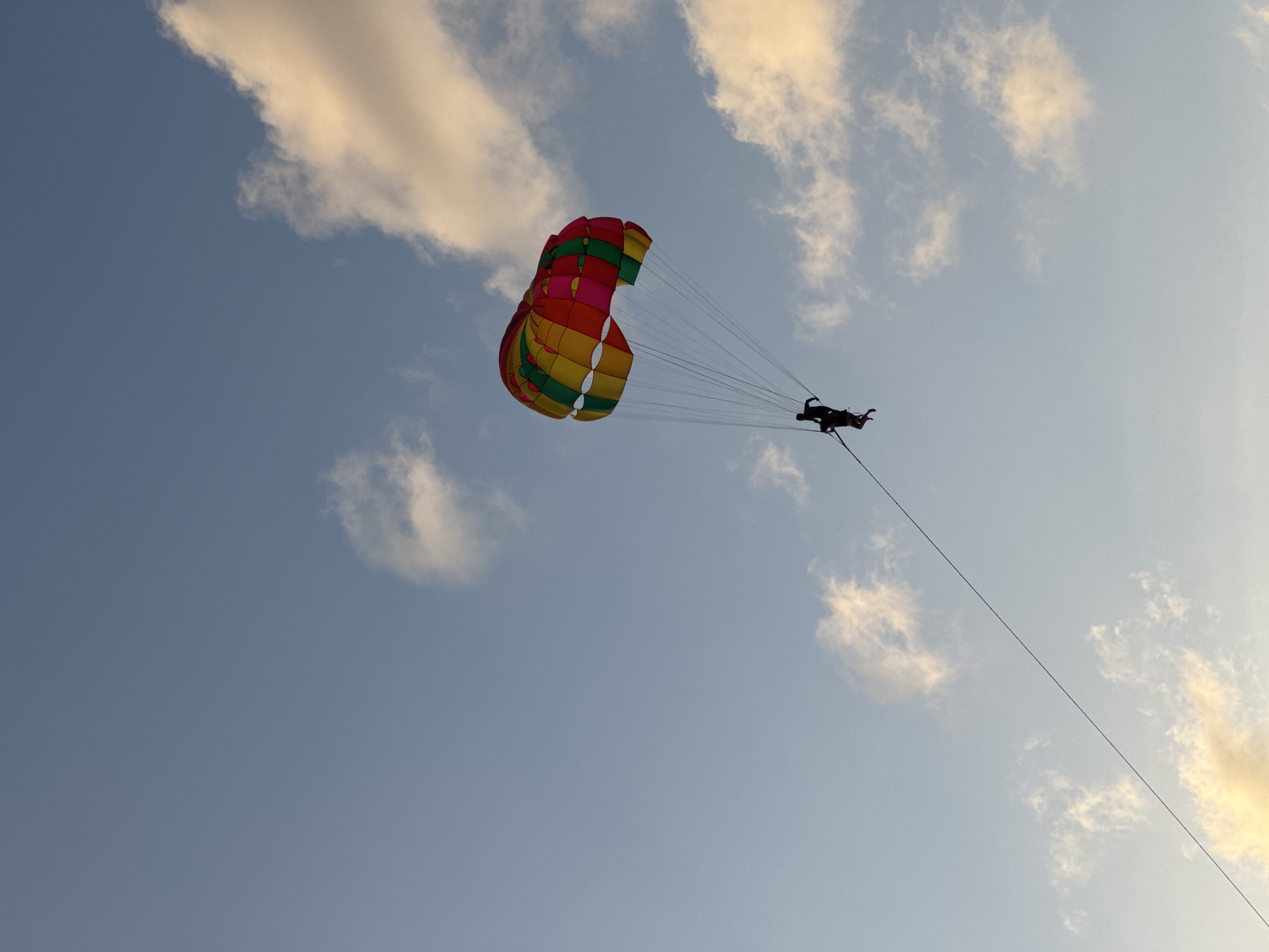A parasailer is being pulled across a light blue sky with scattered clouds by a tow rope, as seen from a low angle. The parasail itself is a large, colorful canopy with alternating red, yellow, and green panels, and it is suspended above two silhouetted figures who are attached to it via harness and ropes. The tow rope extends diagonally downwards to the left, disappearing out of frame. The sky has a gradient of light blue at the top to a slightly warmer tone near the horizon, with some wispy white and golden-tinged clouds. The scene evokes a sense of adventure and leisure, typical of coastal tourist destinations.