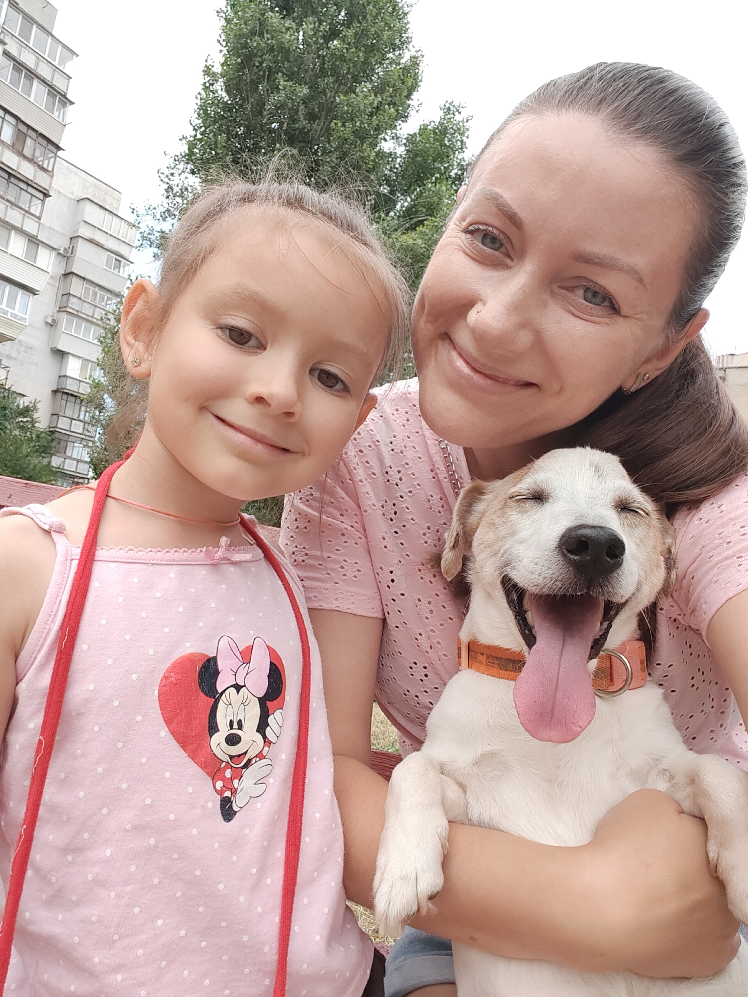 This is a medium shot, outdoor selfie taken in Chornomorsk, Ukraine. A woman and a young girl are in the foreground, with a happy dog between them. The woman, on the right, is smiling and looking at the camera. She has brown hair pulled back and is wearing a pink, short-sleeved shirt. The girl, on the left, is also smiling and looking at the camera. She has brown hair in pigtails and is wearing a pink sleeveless shirt with a Minnie Mouse graphic. The dog, held by the woman, is panting with its tongue out and eyes closed, appearing very joyful. The dog is white with brown markings and is wearing an orange collar. In the background, there is a large apartment building and trees, typical of an urban residential area in Chornomorsk.