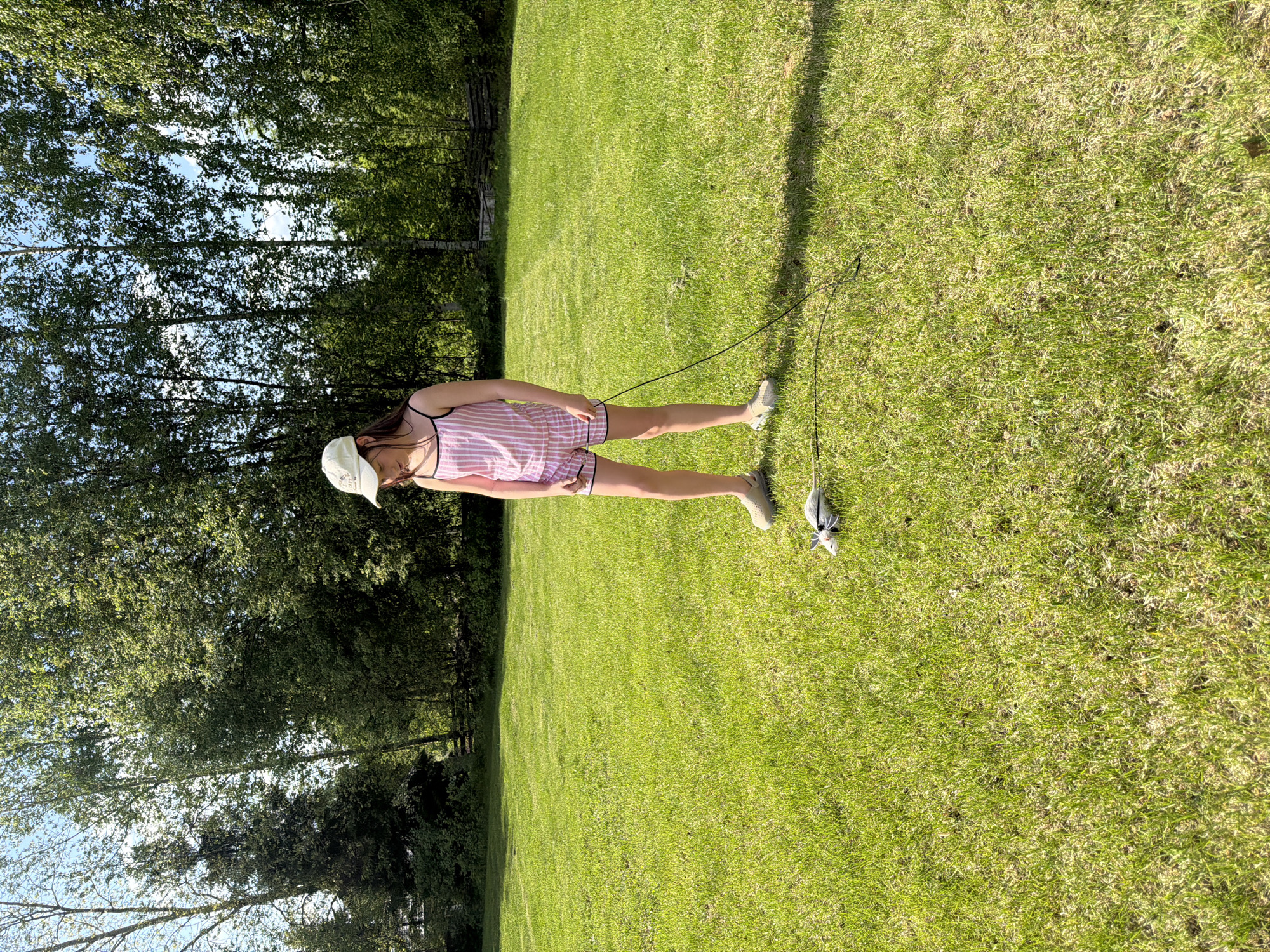 A young woman stands in a grassy field in Dubovaya, Russia. She is wearing a pink and white striped pajama set, a baseball cap, and sneakers. She is holding a string attached to a toy mouse. The background includes trees and a wooden fence.