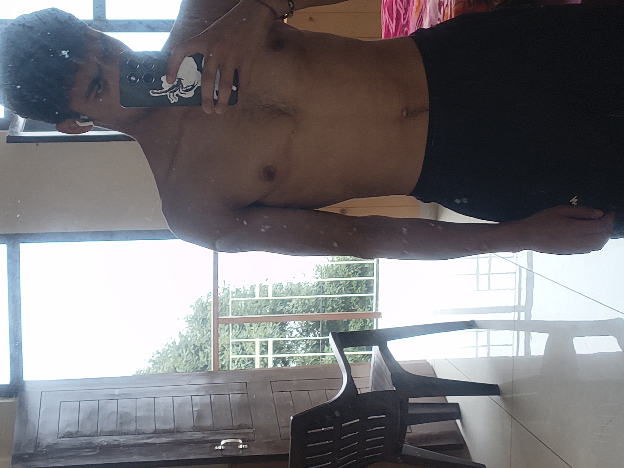 A young man with dark curly hair and bare torso is standing in a room with large windows and a wooden door. He is holding a smartphone in his right hand, taking a selfie. He is wearing black athletic shorts. The room has a tiled floor and a plastic chair on the left. Through the windows, a balcony with a railing and lush green trees can be seen, suggesting an outdoor setting. The lighting in the room is bright, possibly from natural light. There are no other people visible in the image. The image appears to be taken in a residential setting.