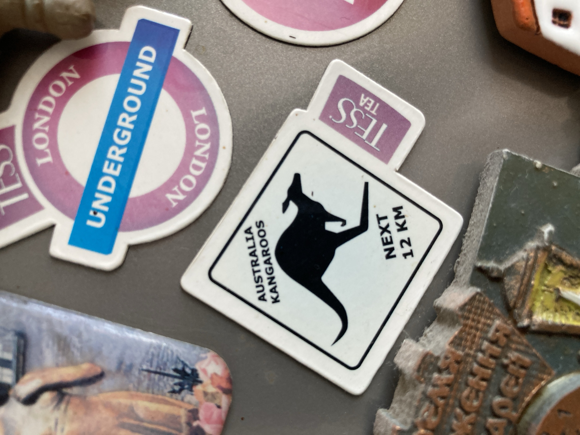 The image shows a collection of refrigerator magnets attached to a metallic surface. The magnets are diverse in design and theme.
Prominently featured is a circular magnet with a purple outer ring and a white inner circle, containing the words "LONDON UNDERGROUND" in blue lettering across the center. The outer ring of this magnet has the word "LONDON" repeated twice, written in a curved fashion.
Next to it, a rectangular magnet displays a black silhouette of a kangaroo in mid-leap, on a white background. Above the kangaroo, in smaller text, it reads "AUSTRALIA KANGAROOS". To the right of the kangaroo, larger text indicates "NEXT 12 KM". This magnet also has a smaller, purple tab extending from its top, with the text "TESS TEA" written vertically.
Another magnet, partially visible, is circular with pink and white coloring, and text that is mostly obscured but appears to be related to London as well.
Further down and to the left, a rectangular magnet is visible with an image of what appears to be a painting of a person or people in a pastoral setting.
To the right, a portion of a textured, metallic-looking magnet is visible, possibly depicting buildings or a city skyline, with some Cyrillic text inscribed on it. The overall scene is a close-up shot of refrigerator magnets, suggesting a casual domestic setting. The lighting appears to be ambient, with no strong shadows, implying an indoor environment.