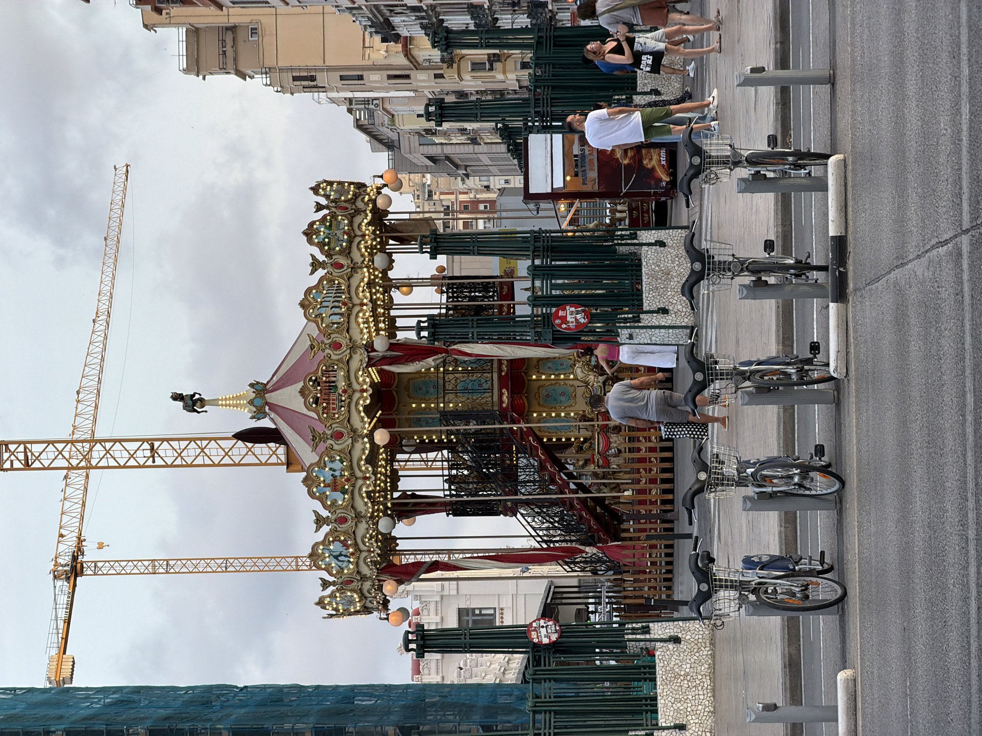 A carousel is on display in an urban area. The carousel has a red and white striped roof and is decorated with lights and ornamental figures. Behind the carousel, a construction crane is visible. In the foreground, several rental bicycles are parked. People are seen walking in the background. The scene appears to be set in a city, possibly during the day, given the lighting conditions. The architecture of the surrounding buildings suggests an urban environment.