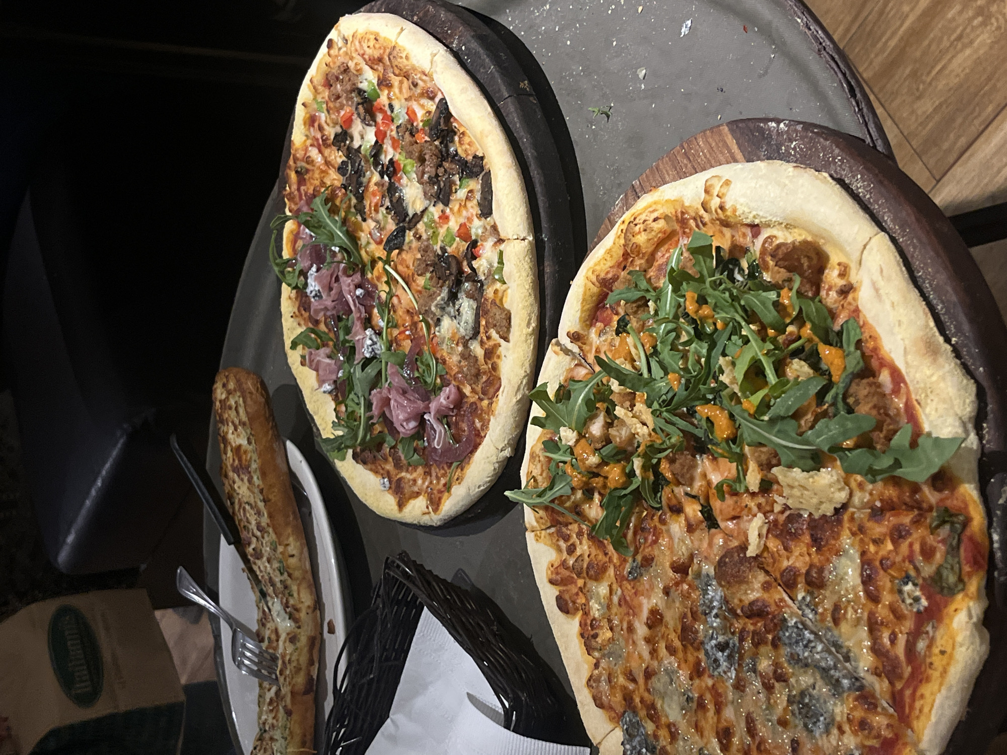 The image shows two pizzas and a bread loaf on a table. The pizza on the left has toppings of vegetables, pepperoni, sausage, and arugula on a wooden circular tray on top of a dark metal table. The pizza on the right has toppings of cheese, chicken, hot sauce, and arugula, and it also sits on a wooden circular tray on top of a dark metal table. A plate containing a large loaf of garlic bread with a fork and knife is on the left side of the table. A black basket with a white napkin is between the bread plate and the pizzas. In the lower left corner, there is a bag with the word "Italianis" printed on it.