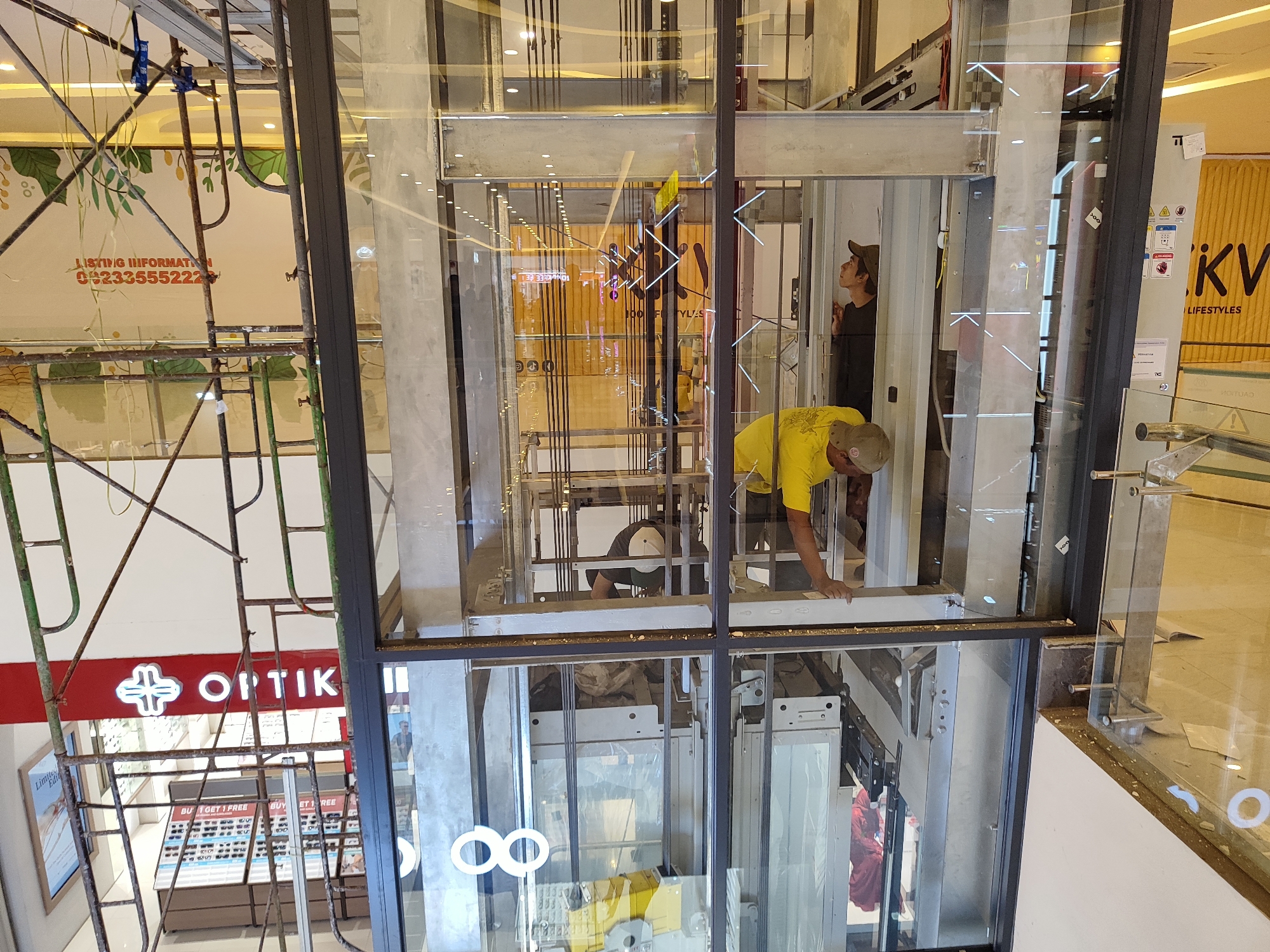 This is a medium shot, eye-level, indoor photograph of an elevator shaft under construction in a commercial building. The elevator shaft is enclosed by glass on at least one side, and scaffolding is visible in the foreground, obscuring some of the view. Inside the shaft, there are two men, one wearing a yellow shirt and the other wearing a dark shirt and a cap. The man in the yellow shirt appears to be working on the elevator mechanism, while the other man is looking up. The elevator shaft is surrounded by concrete walls and metal framing. In the background, a sign for "OPTIK" and a display of glasses are visible through the glass. The lighting appears to be artificial, emanating from overhead fixtures and possibly from lights within the elevator shaft. The overall impression is one of ongoing construction and renovation work. The image is undated and unlocated, but it depicts a scene that could be found in any urban or commercial area. It is noted that the scene is located in Sempusari, Indonesia.