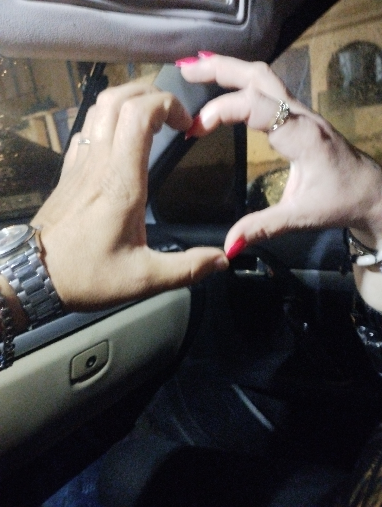 Two hands, one with a watch and the other with bracelets, are forming a heart shape inside a car. One hand has pink nail polish, the other red. The hands are adorned with rings. The interior of the car and a building outside the car window suggest that the photo was taken in Martil ⵎⴰⵔْتِيل مرتيل, Morocco.