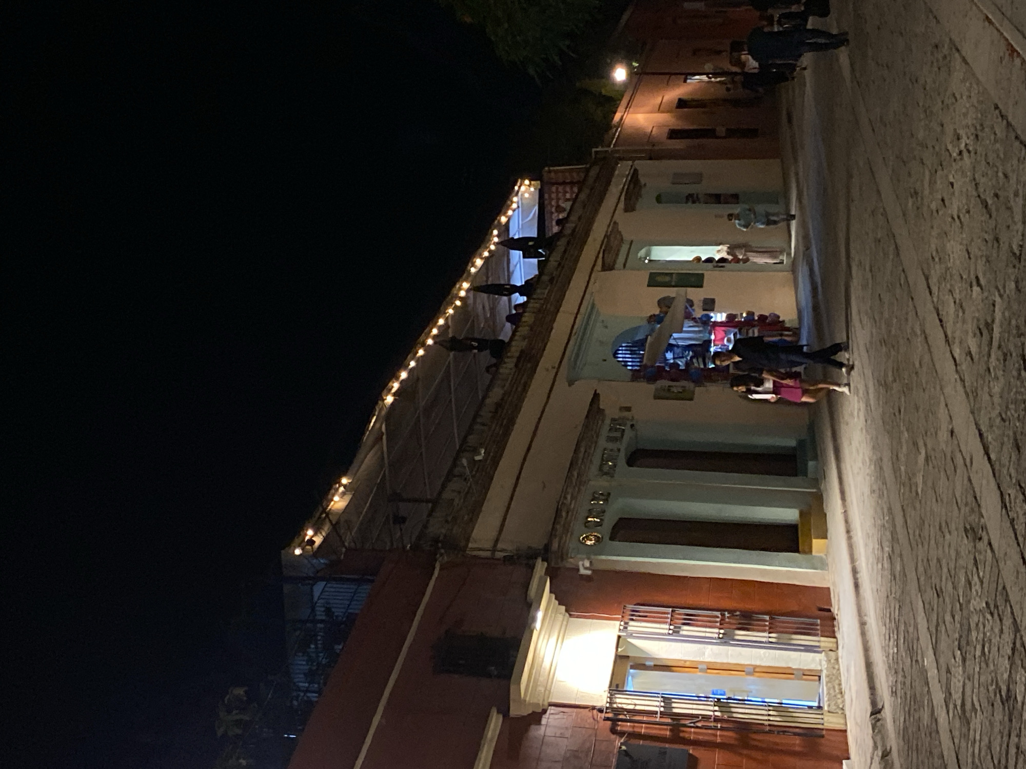 At night in Oaxaca City, Mexico, a building with a balcony lit by string lights is seen. Silhouetted figures stand on the balcony. The building's facade, with windows and columns, faces a street where people are walking. The street is paved with cobblestones. The sky above is dark.