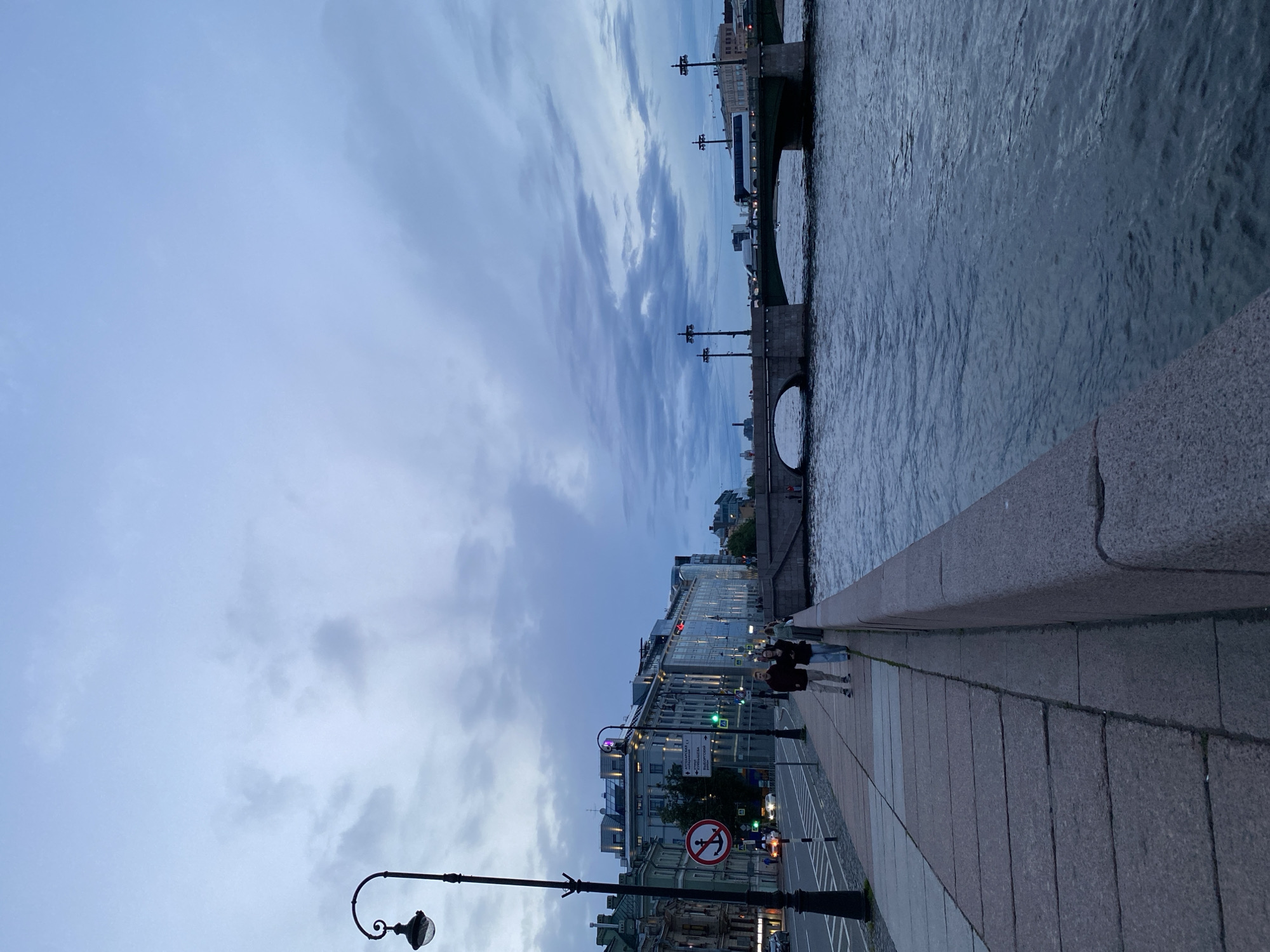 The image depicts a waterfront scene in Saint Petersburg, Russia, likely in the evening given the twilight sky. The sky is filled with dramatic, wispy clouds in shades of grey and pale blue, suggesting overcast or nearing-stormy weather. A wide river, with choppy, dark water reflecting the muted sky, occupies the right side of the frame.
A granite embankment runs along the river, creating a walkway. On this walkway, a few people are visible, appearing as silhouetted figures walking or standing. Two individuals are particularly noticeable, dressed casually and walking away from the camera. Further down the embankment, more figures are spread out.
Adjacent to the embankment is a city street lined with multi-story buildings. These buildings exhibit a mix of architectural styles, with some appearing more modern and glass-fronted, while others have a more traditional, ornate facade. Streetlights and lampposts, one with an ornate, curved design, are interspersed along the sidewalk and street. Traffic lights and street signs are also visible, including a circular red and white sign with an anchor symbol, indicating a prohibition, likely related to boating or navigation. A few cars are on the street, their headlights casting a faint glow.
In the distance, a bridge spans the river, with structural elements and lampposts visible against the sky. The overall mood is atmospheric and somewhat somber, with the fading light and cloudy sky contributing to a sense of quiet introspection. The scene captures a moment of urban life along a historic waterway.