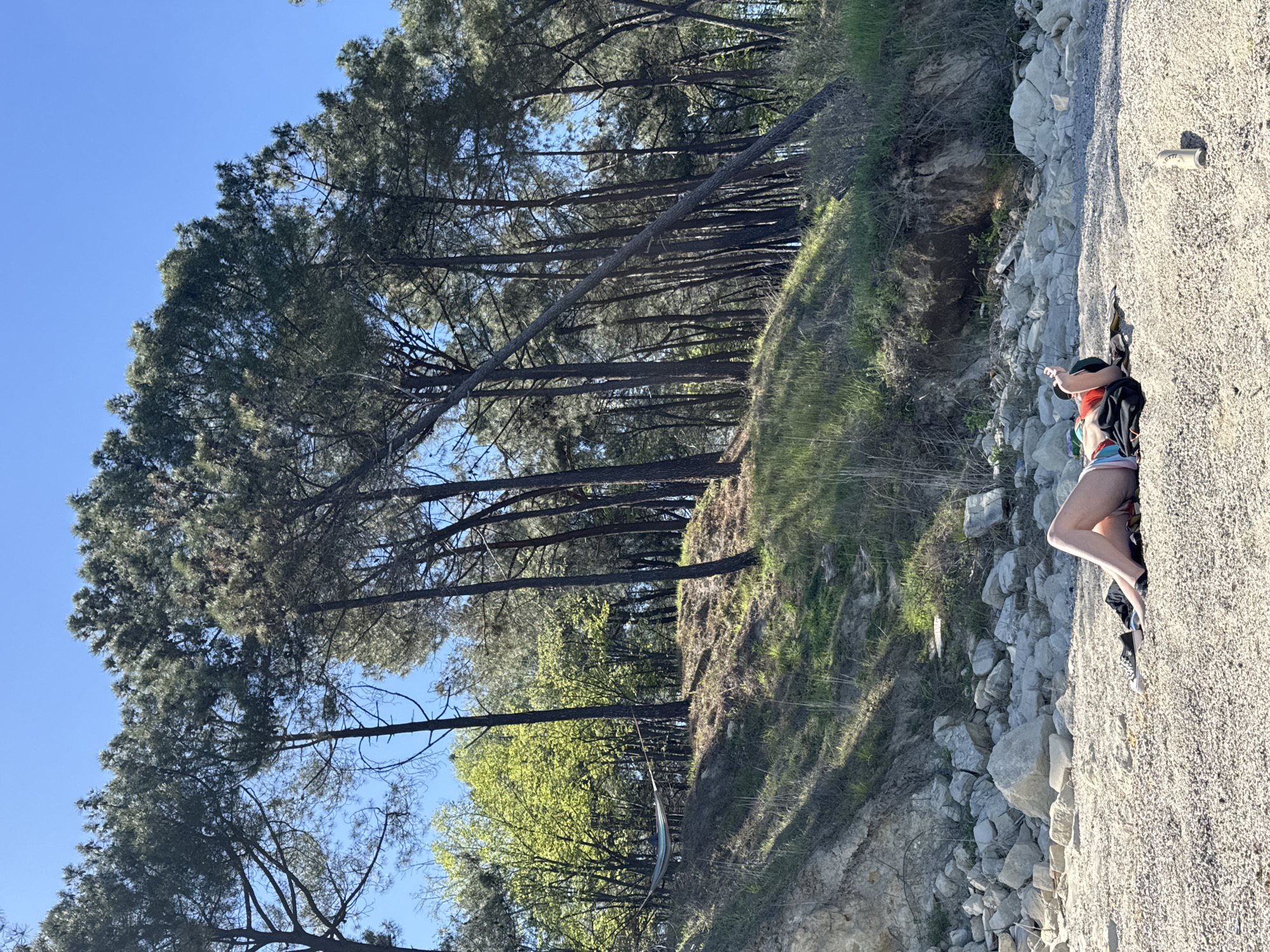 A medium, rotated shot shows a woman on a gravelly path in Varna, Bulgaria. She wears a red hat, dark shirt, shorts, and backpack. A slope descends to a wooded area. Tall trees stand on the slope. The sky is blue.