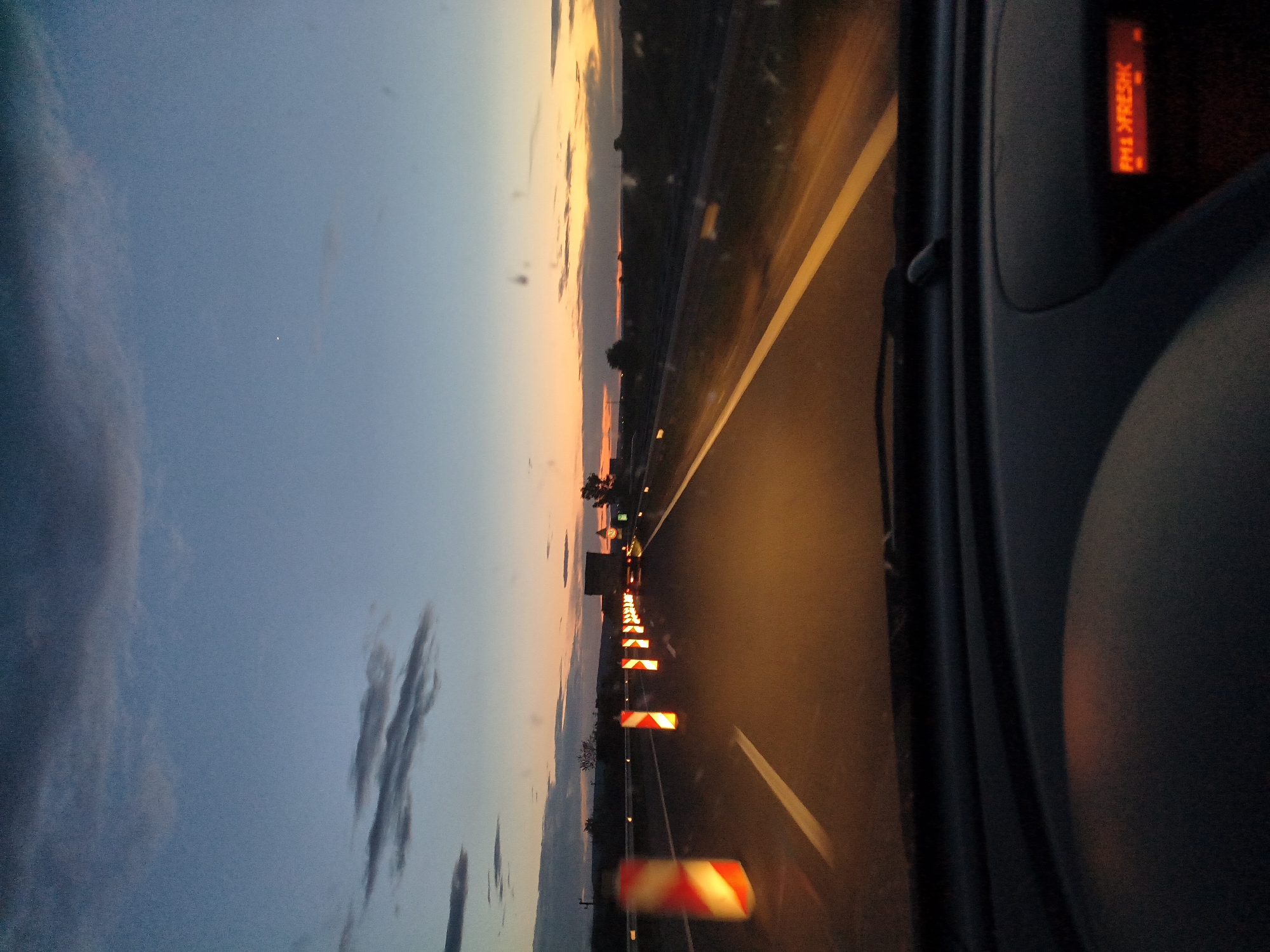 The image depicts a view from inside a vehicle, likely on a highway near Shumen, Bulgaria. The view is from the driver's perspective, looking out onto a road with yellow lane markers. Traffic cones and warning signs line the side of the road. The sky is visible with hues of orange at the horizon. The vehicle's dashboard or side panel is visible on the right, with a red text display. The text reads "PROSZEK" in bright red capital letters. The time of day is likely twilight or dusk. There are no people visible in the image.