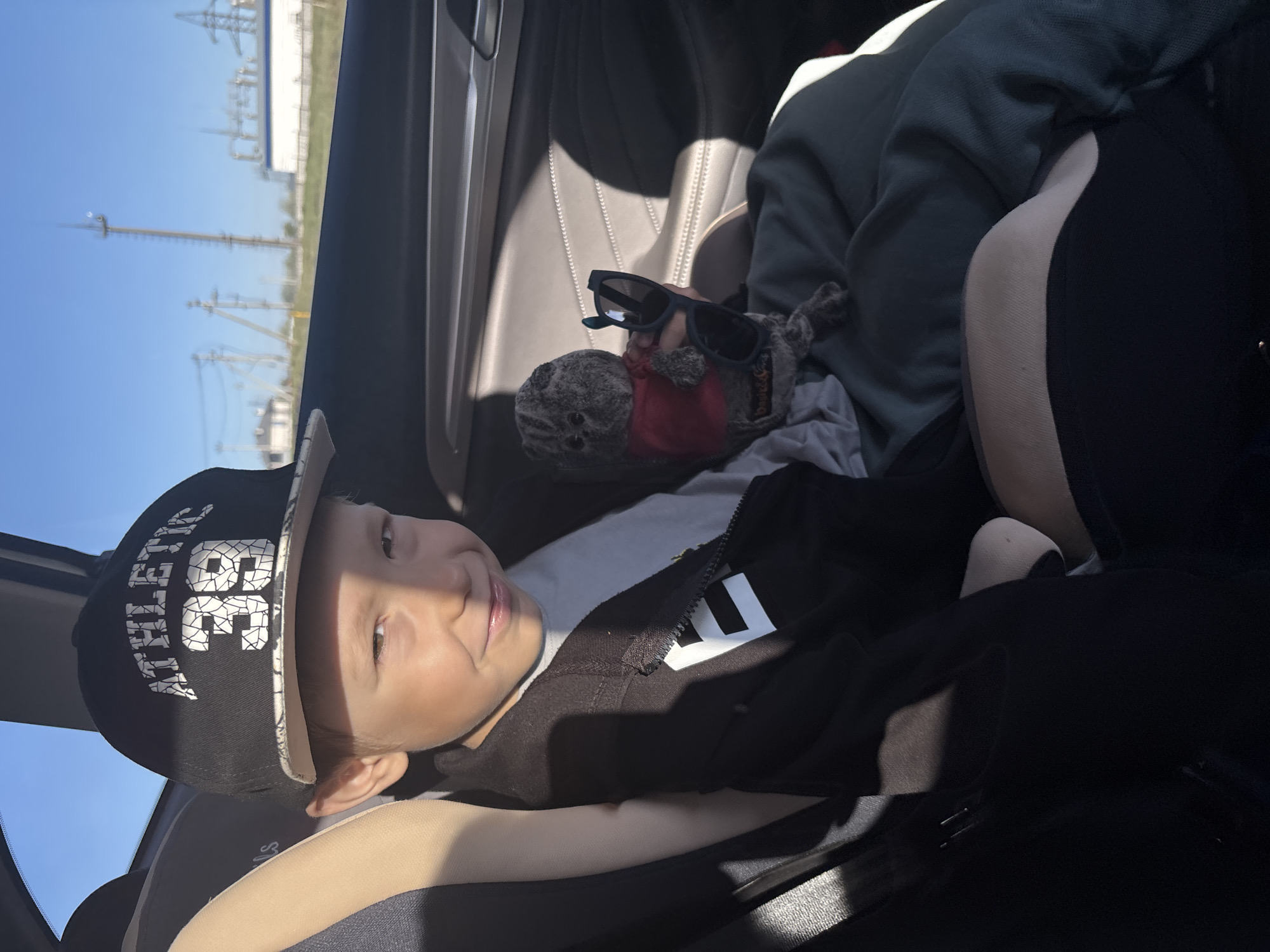 A young child wearing a black baseball cap and black jacket reclines in a car seat. A stuffed animal wearing sunglasses is situated beside the child. Outside the car window, a blurred view of what seems to be the edge of Chaikovsky, Russia, shows utility poles and buildings. The sky is blue. The car's interior is tan and black.
