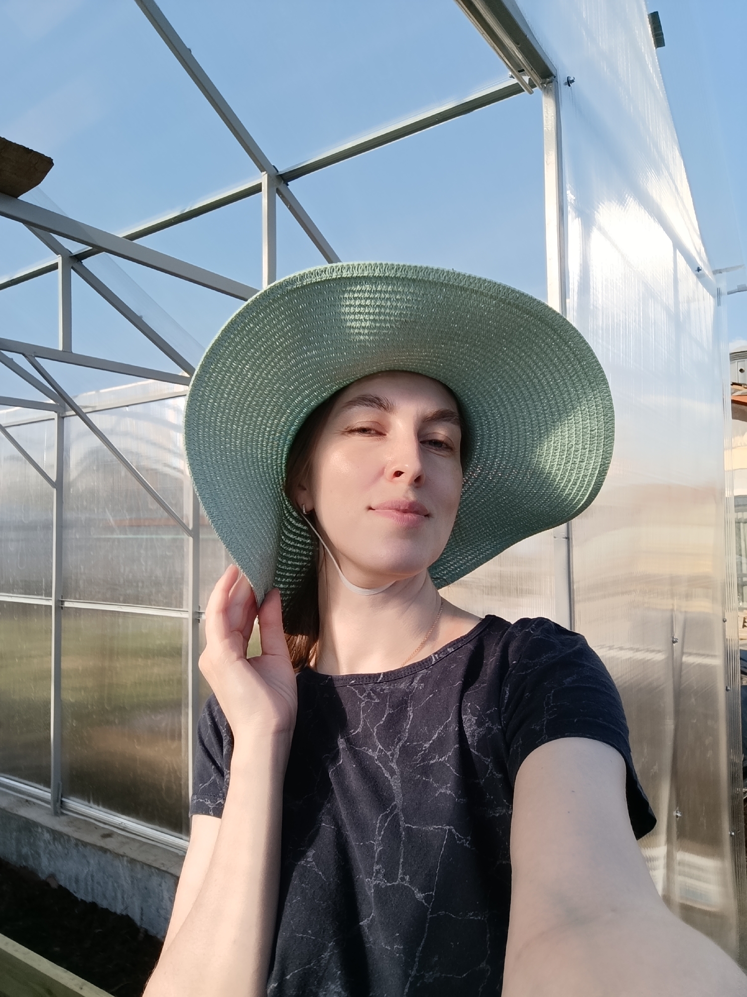 A woman with brown hair wearing a wide-brimmed, light-green hat and a dark-colored t-shirt with a marbled pattern poses for a selfie. She is holding the brim of her hat with her right hand. Behind her is a structure that appears to be a greenhouse or similar outdoor enclosure with a metal frame and translucent panels. The location is Chaikovsky, Russia. The sky is clear and blue.