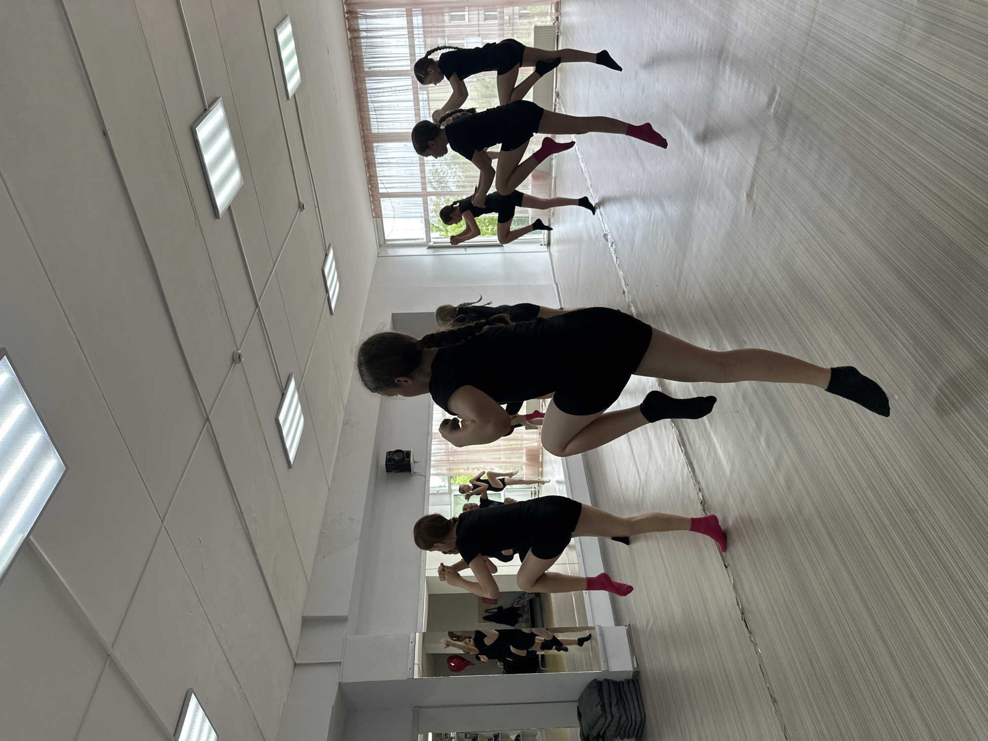 In Votkinsk, Russia, a dance class is in session within a studio. Seven young girls, dressed in black leotards and shorts, are practicing a dance routine. Their movements suggest a combination of ballet and martial arts, with one leg raised in front of the body, bent at the knee. Some wear pink socks, while others wear black. The studio features a large mirror along one wall, reflecting the dancers and the room's interior, including fluorescent ceiling lights and large windows with sheer curtains. The floor is a light wood-grain laminate. The environment appears clean and functional, typical of a rehearsal space.